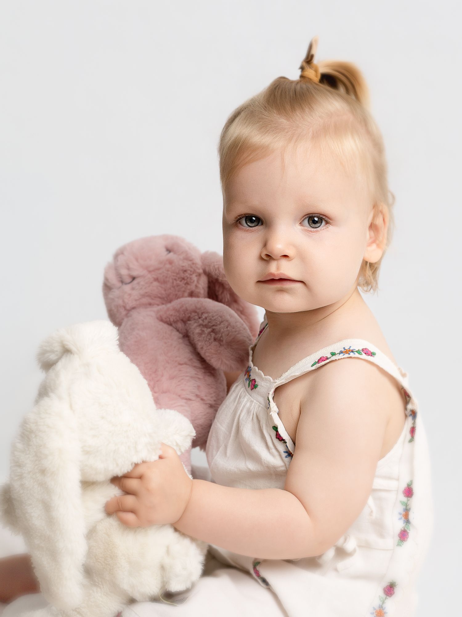 Toddler girl with blonde hair holding white and pink stuffed animal toys against white background.