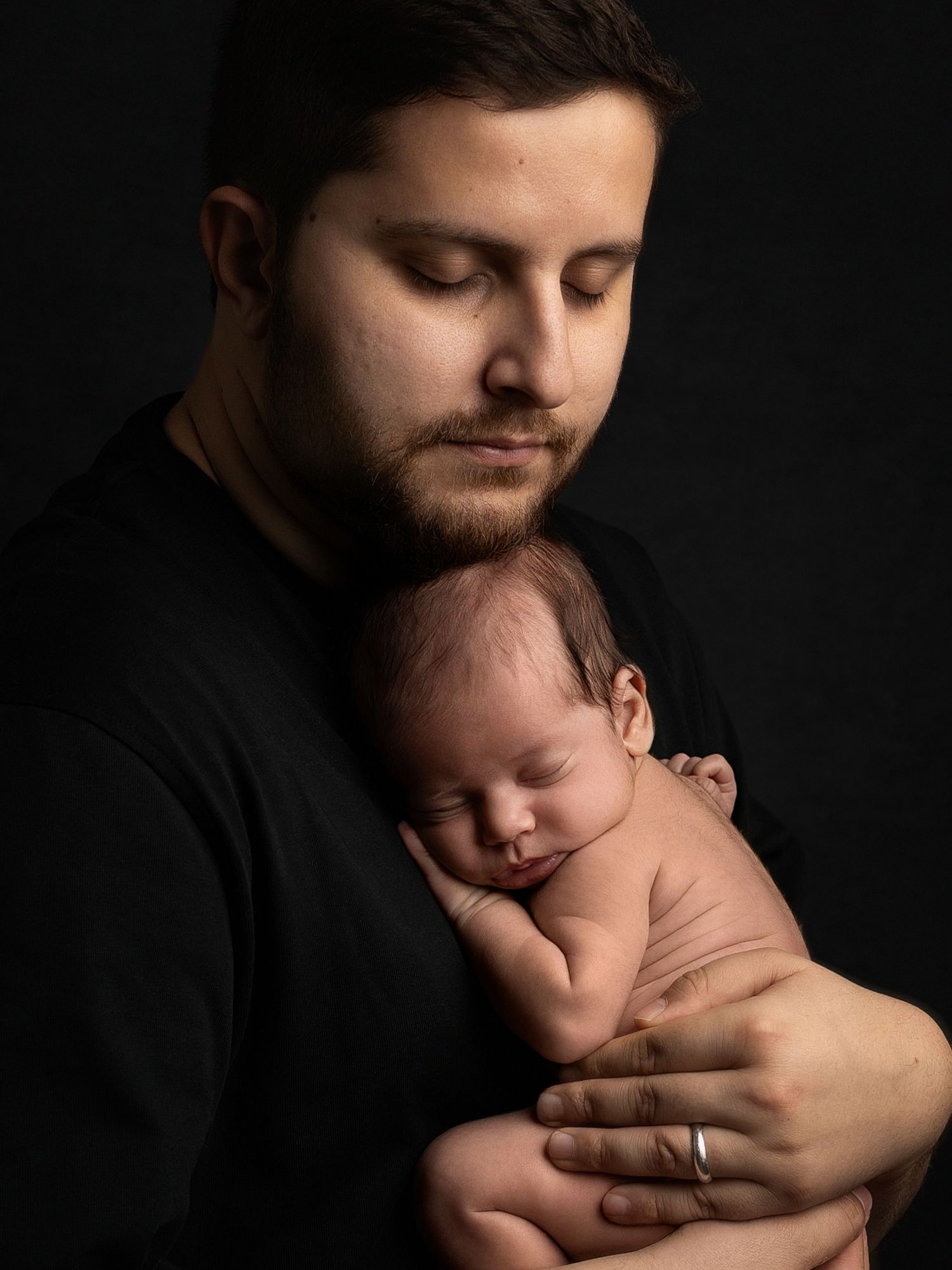 A father in a black shirt tenderly holds his sleeping newborn baby against his chest in a studio portrait.