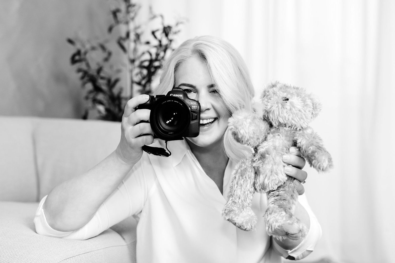 Smiling blonde woman holding a DSLR camera and teddy bear in a bright, elegant room, black and white photo.
