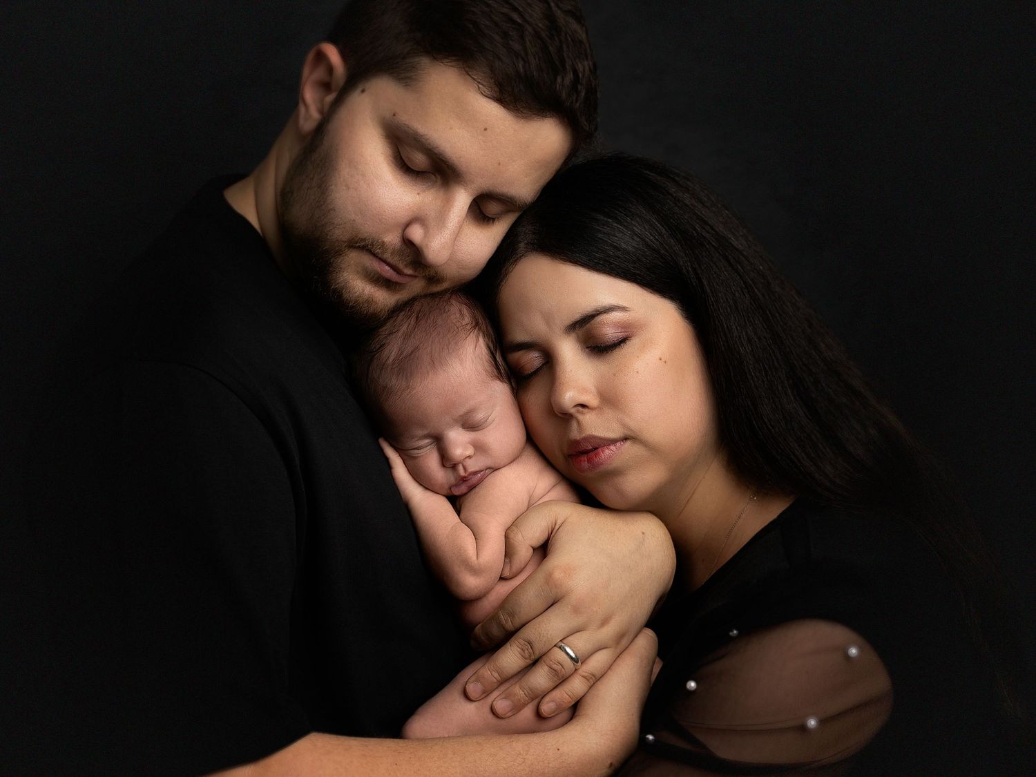 Parents in black clothing tenderly hold their sleeping newborn baby in a professional studio portrait.