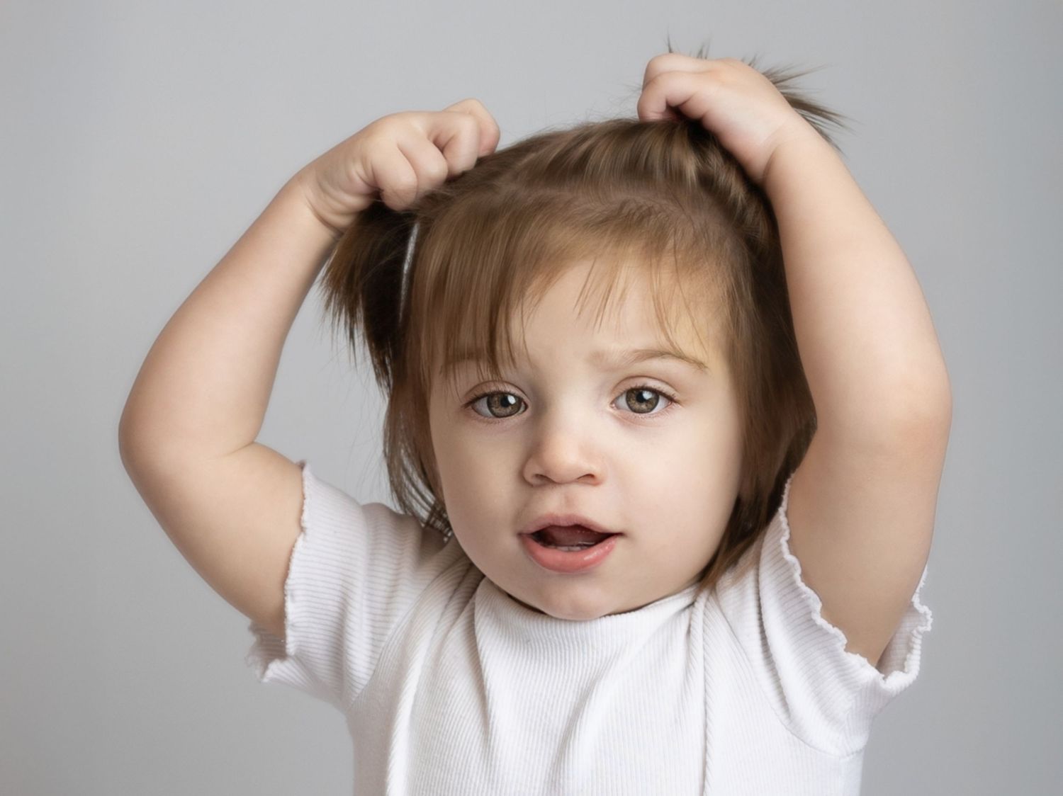 Cute toddler girl in white shirt pulling her hair up with both hands, making a funny face against gray background.