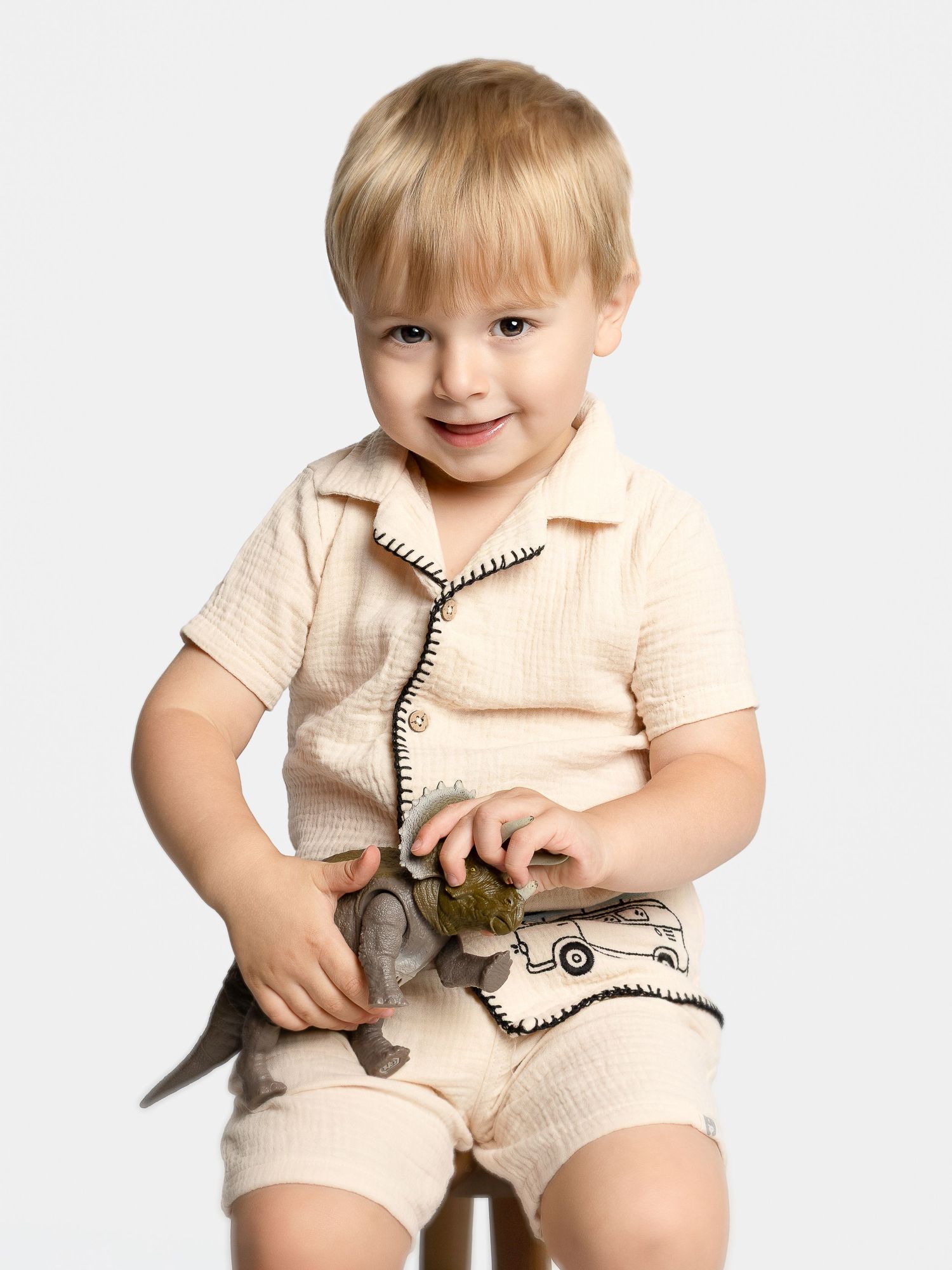 Smiling toddler boy in beige textured short set holding a toy, seated on a stool against a white background.
