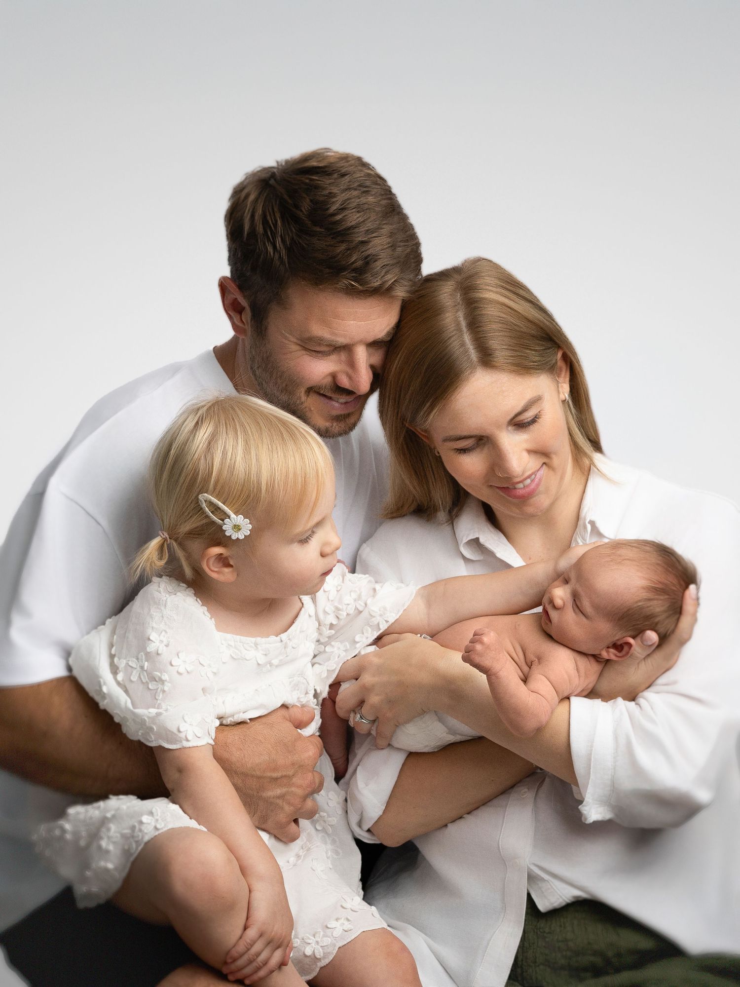 Happy family portrait with parents in white holding a newborn baby and a toddler girl in a white dress.