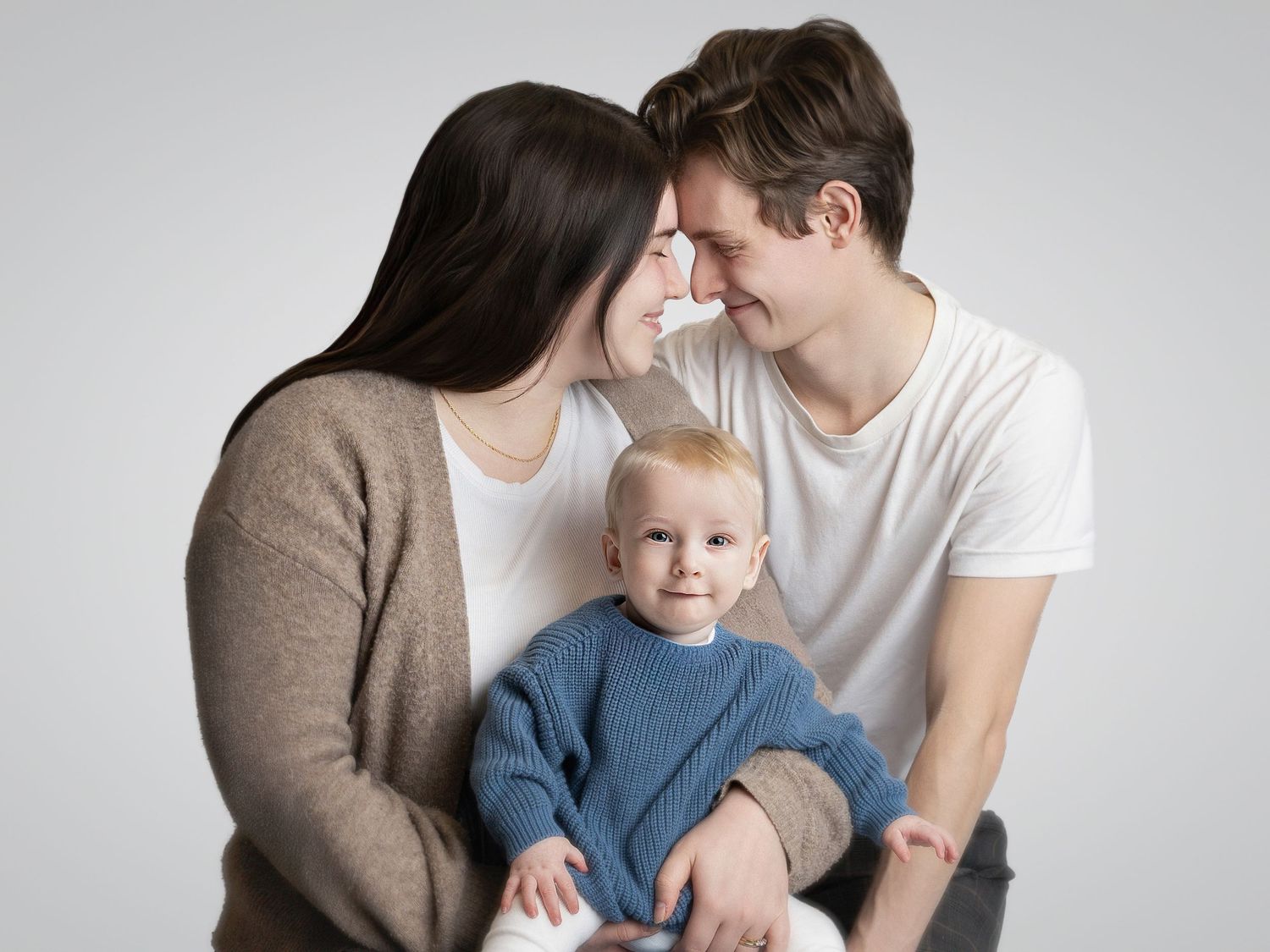 Young family portrait with mother and father smiling at each other while holding their blonde baby boy in a blue sweater.