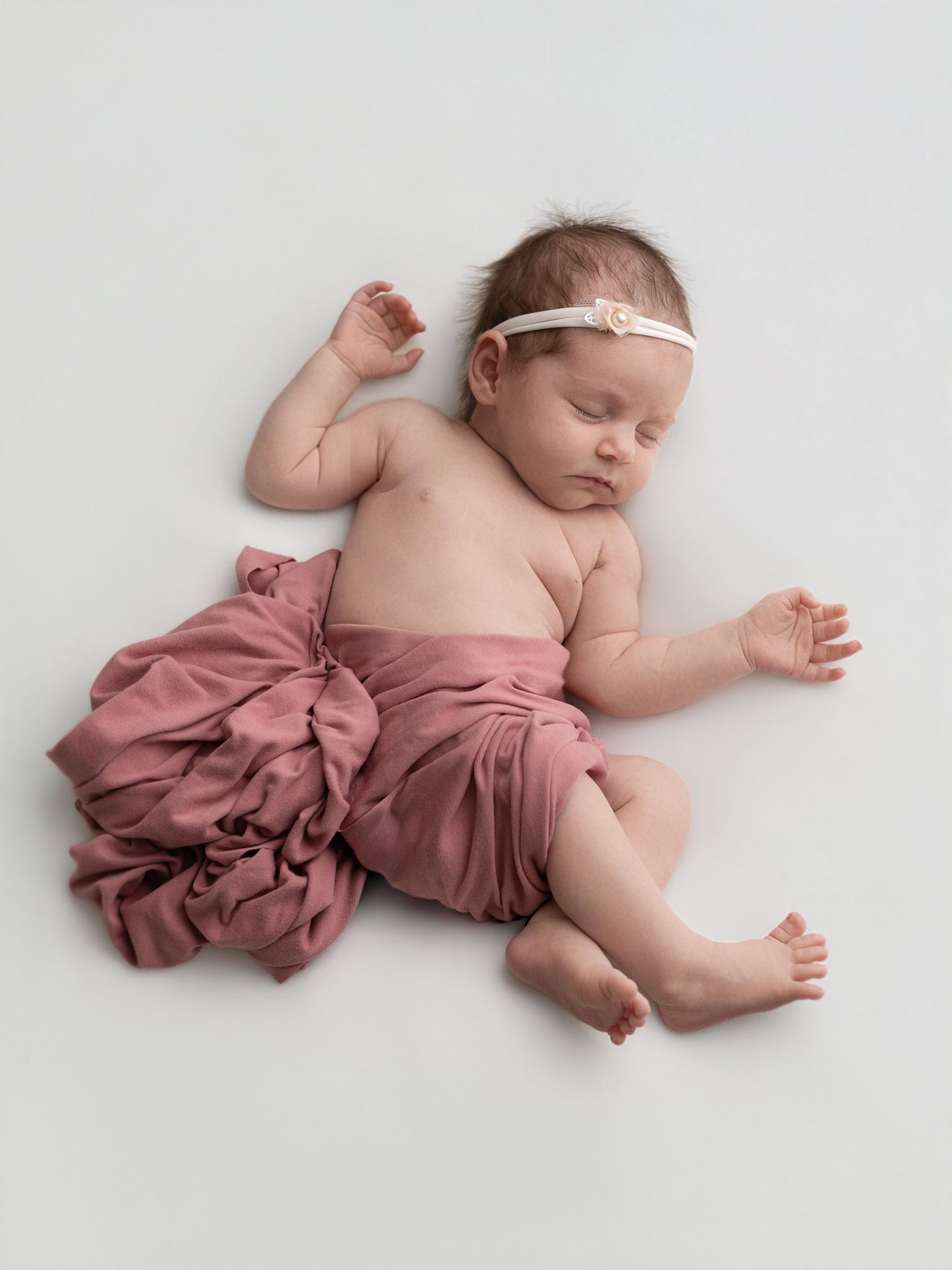 Sleeping newborn baby girl wearing a pink ruffled wrap and white floral headband on a white background.