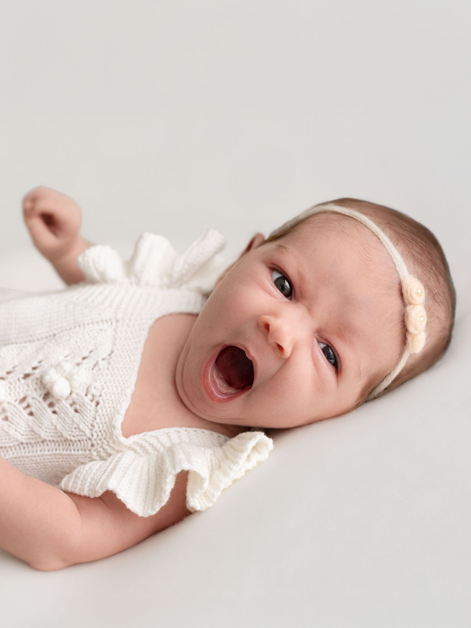 A newborn baby girl in a white knit romper and headband yawns cutely while lying on a white background.