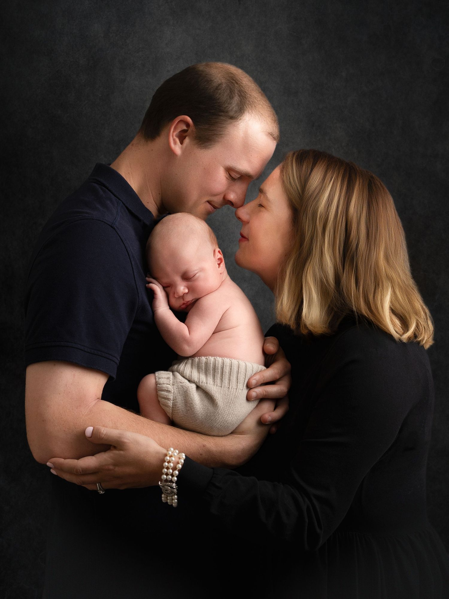 Parents tenderly touch foreheads while cradling their sleeping newborn baby in a professional studio portrait.