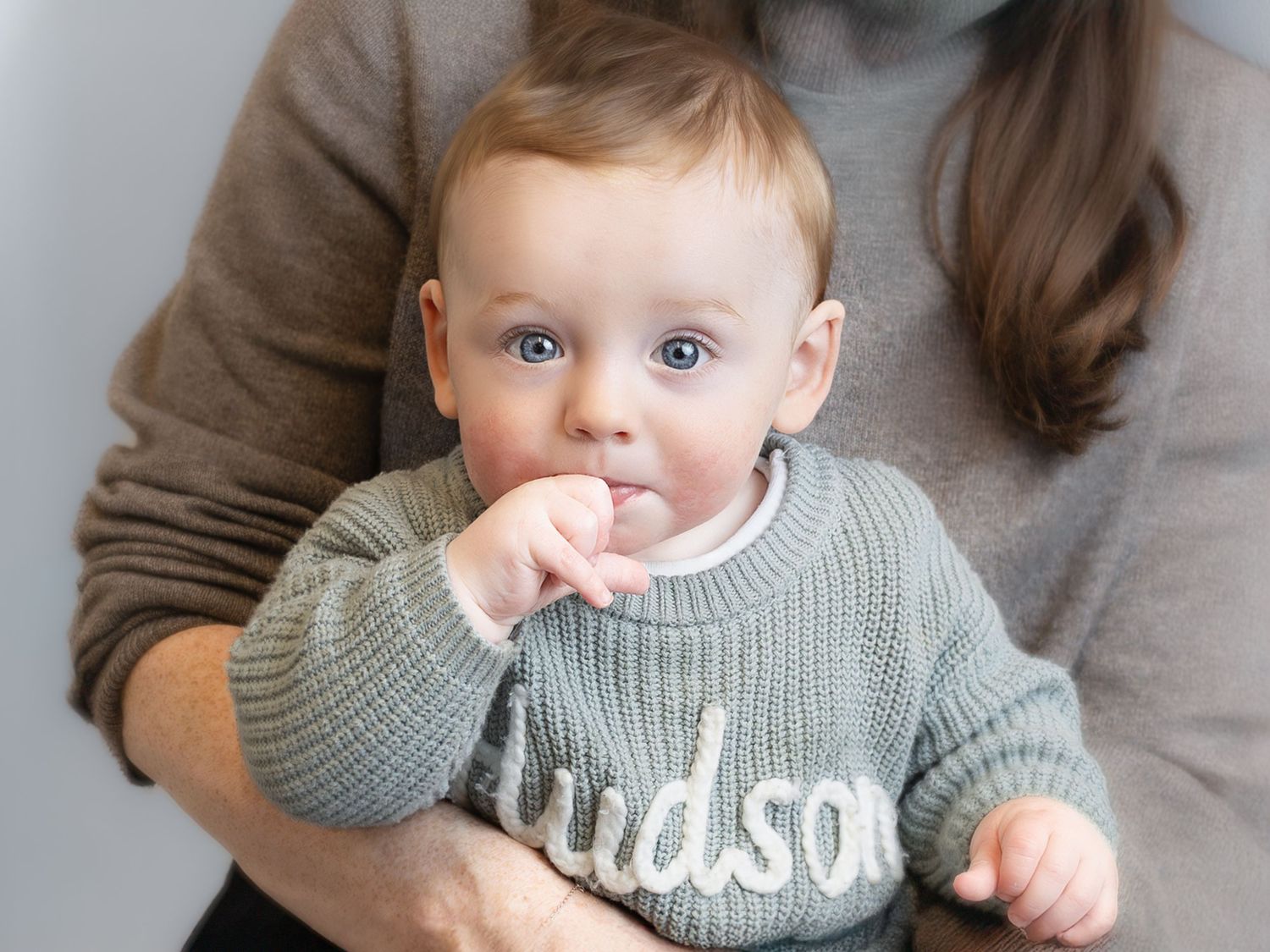 Baby in a sage green knit sweater with the name Hudson sits in the arms of a woman wearing a gray sweater.