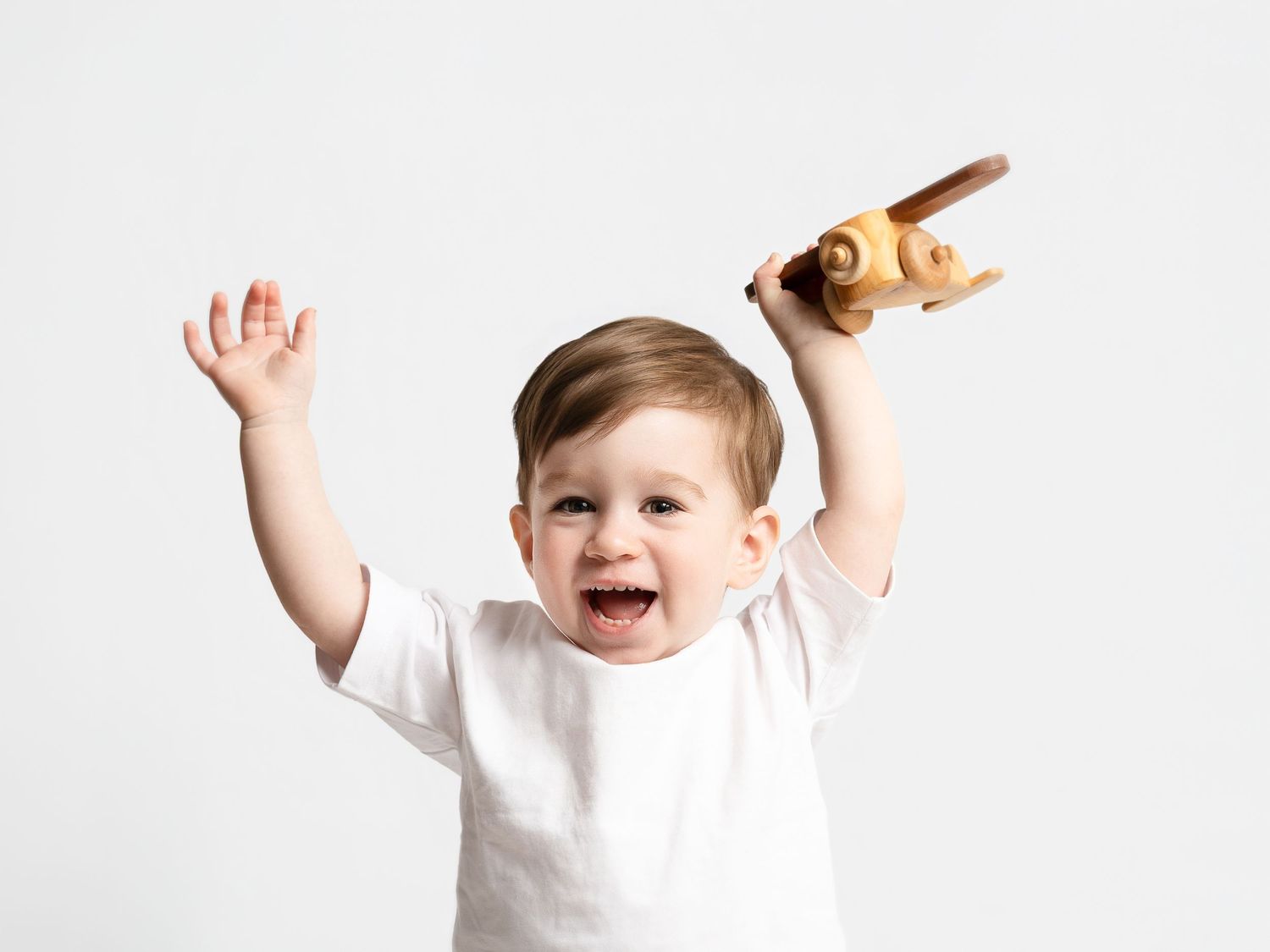 Happy toddler in white shirt holding wooden toy airplane with arms raised against white background.