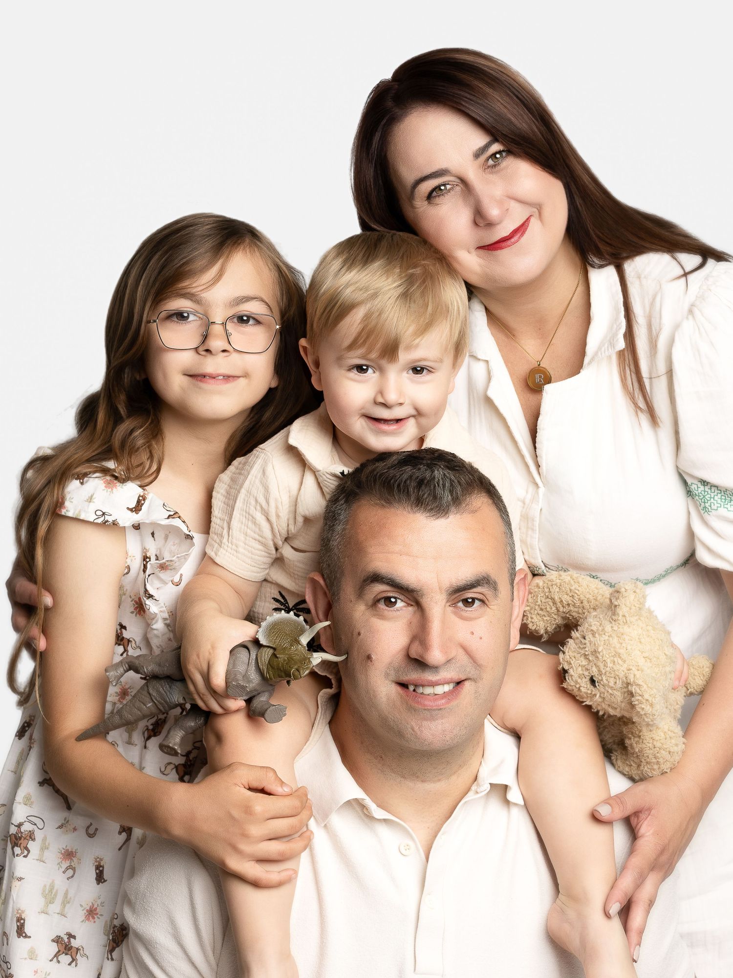 A happy family of four posing for a portrait, parents with two young children holding a teddy bear and toy dinosaur.