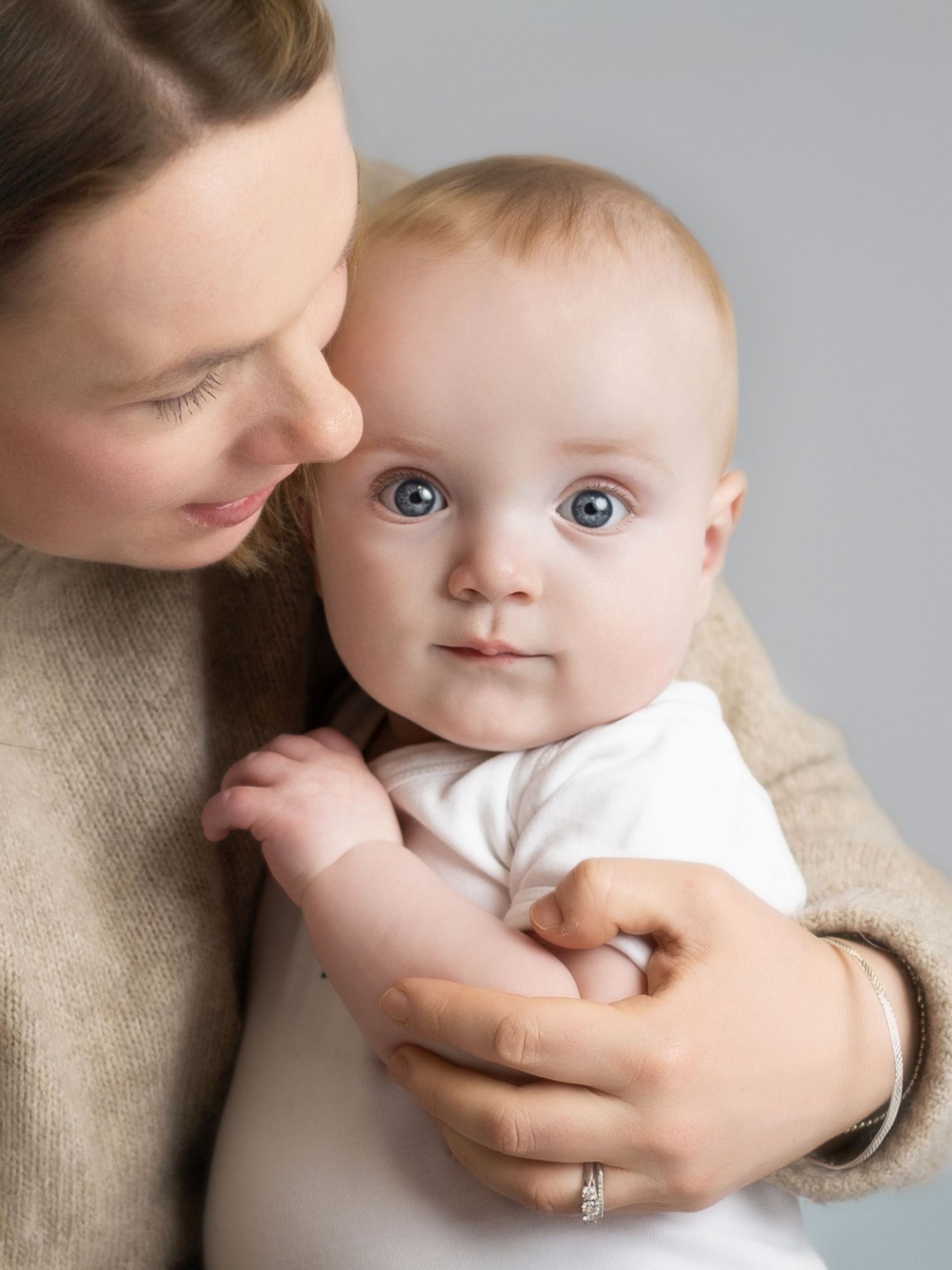 A mother tenderly holds her blue-eyed baby in a white onesie against a soft gray background.