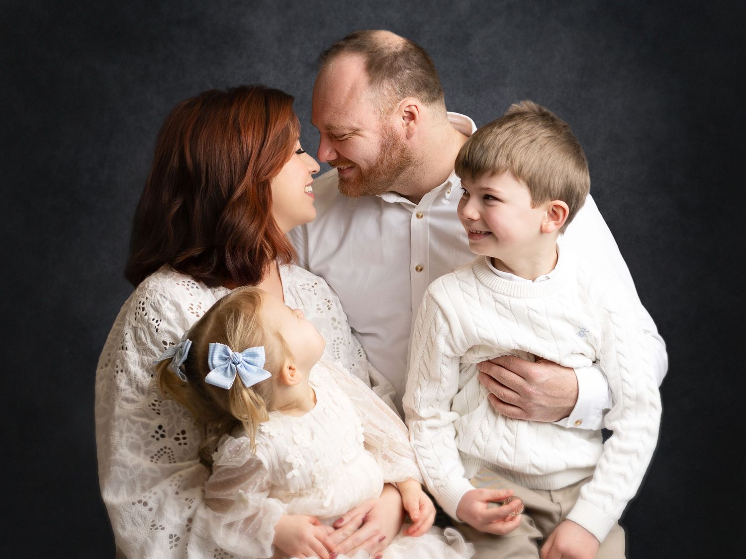Family portrait of parents with toddler girl and young boy, all dressed in white, smiling together on dark background.