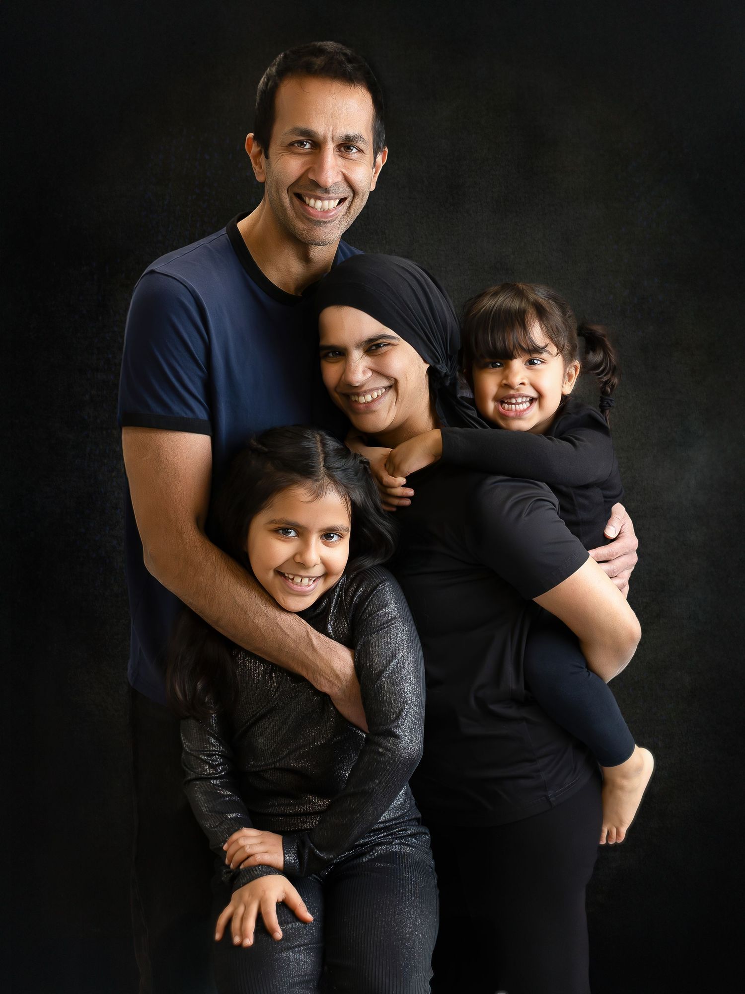 A family of four wearing dark clothing poses together against a black background for a professional studio portrait.