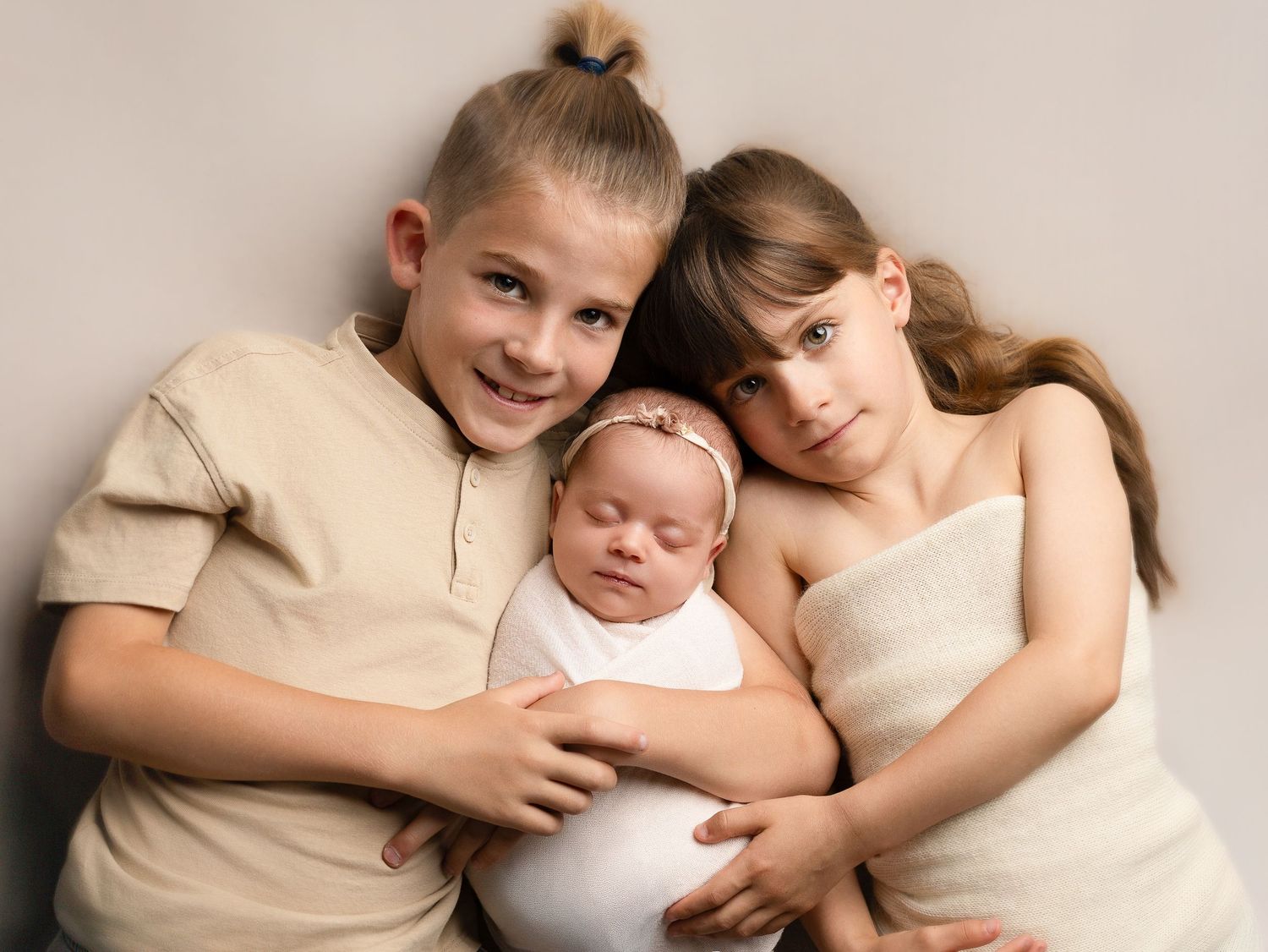 Two children tenderly holding a sleeping newborn baby wrapped in white, posed together in a soft studio portrait.