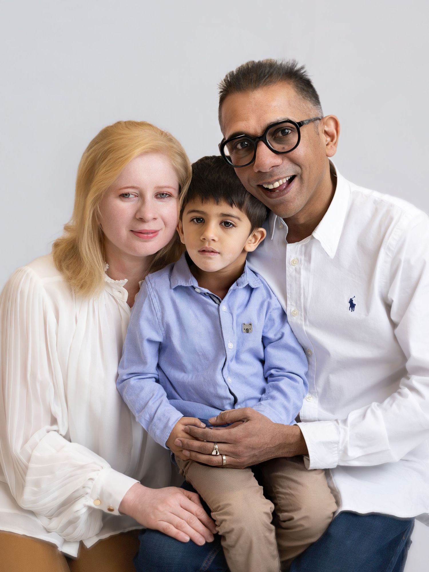 A smiling family of three poses together, with a woman, young boy, and man in glasses wearing white shirts.