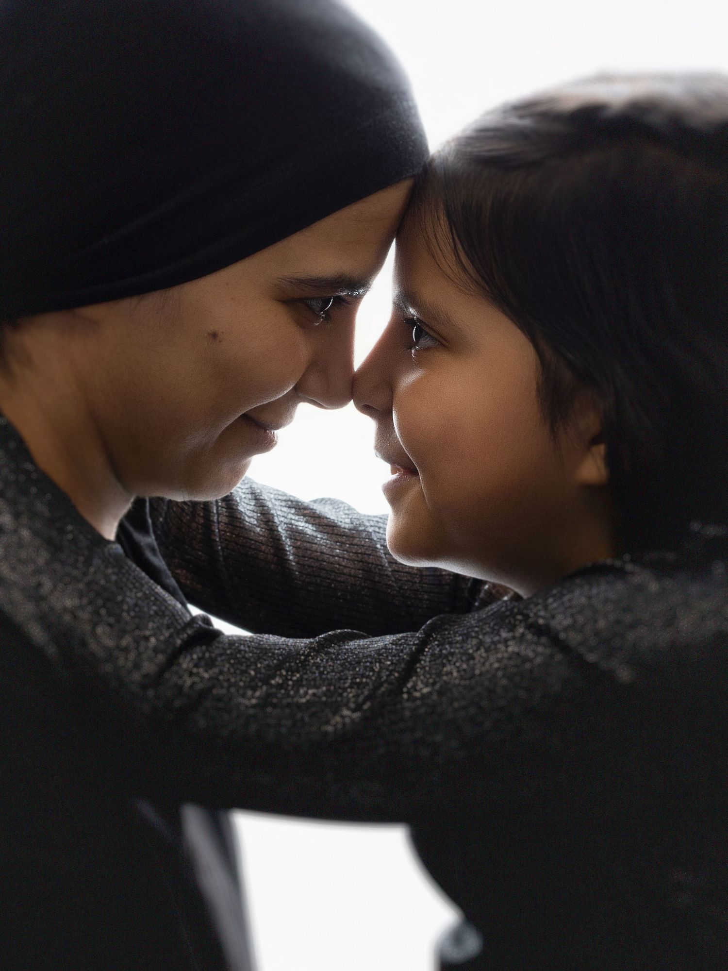 A woman in a hijab tenderly touches foreheads with a smiling child in an intimate, loving moment.