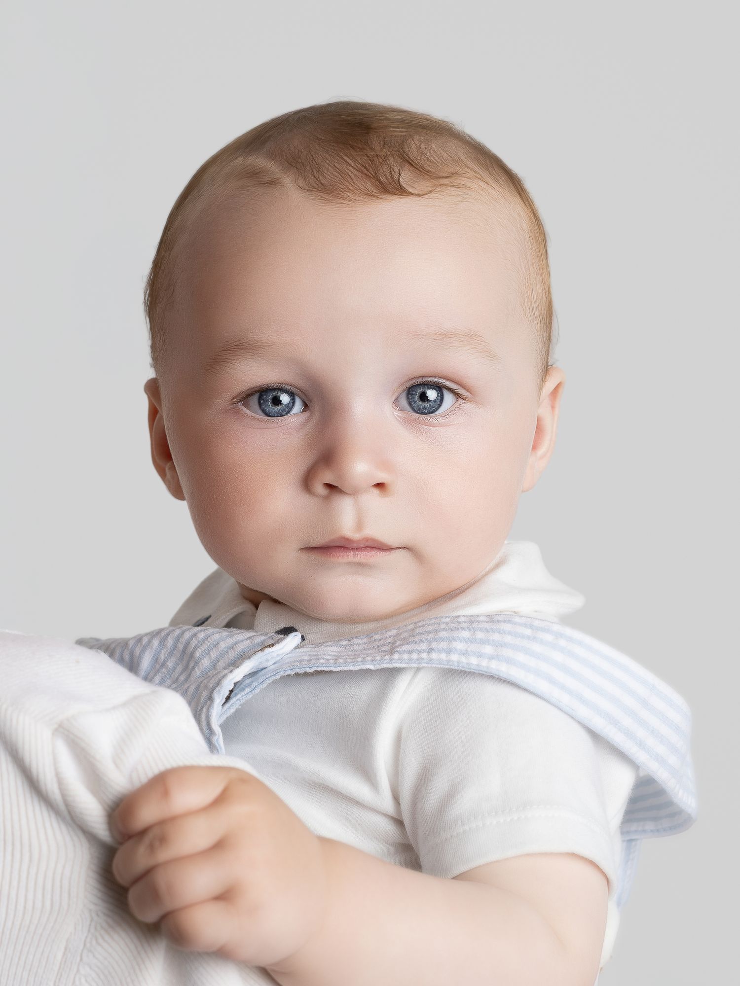A cute baby with blue eyes wearing a white onesie and striped collar, looking directly at the camera.