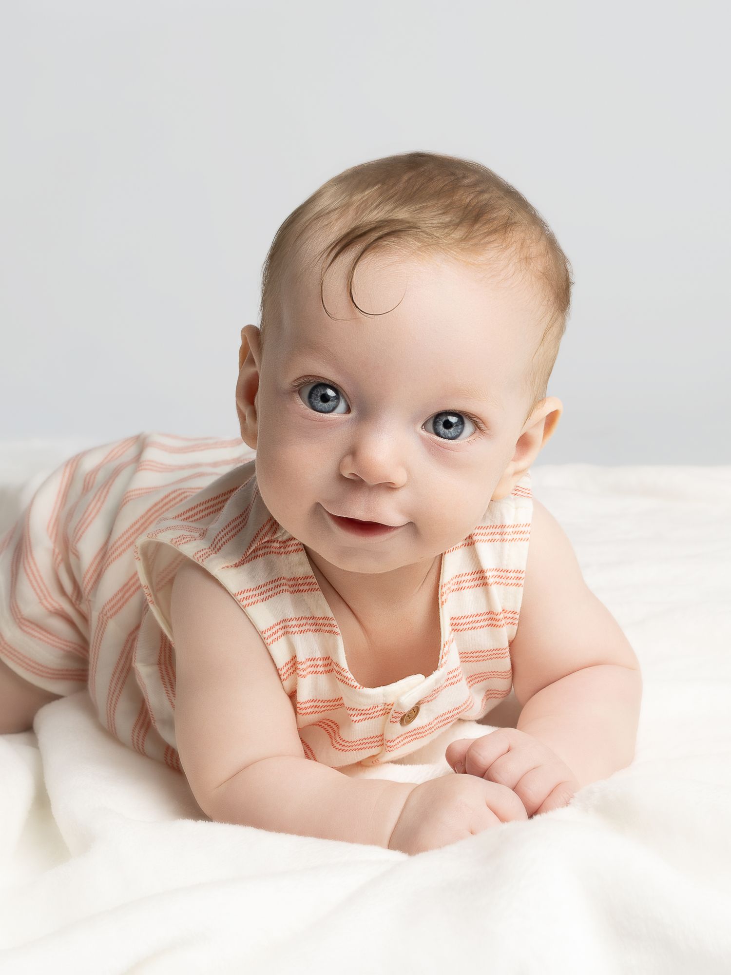 Smiling baby with blue eyes wearing a striped onesie, lying on tummy on a white blanket.