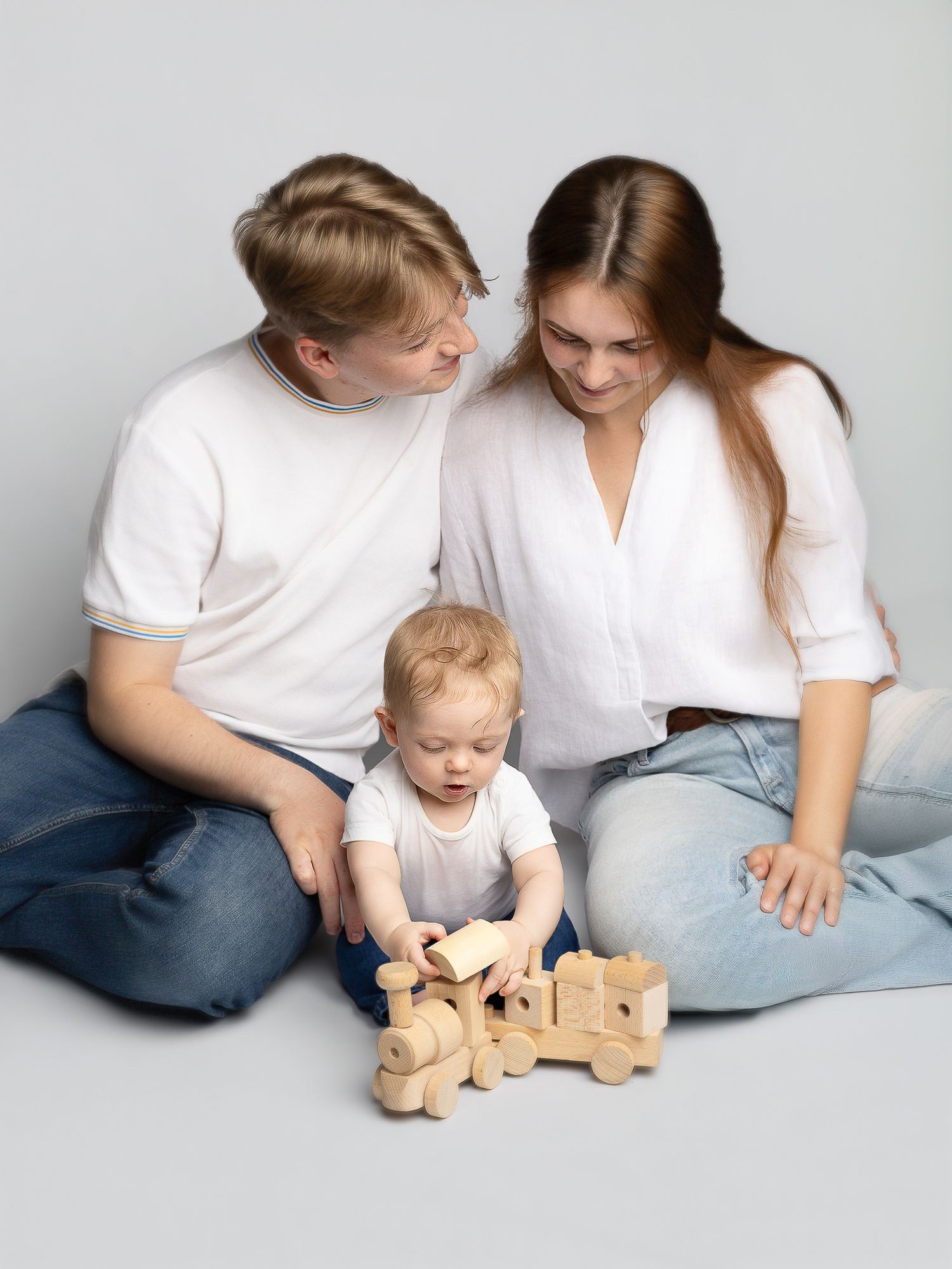 Young family with baby playing with wooden toy train, parents smiling, white shirts and jeans, light background.