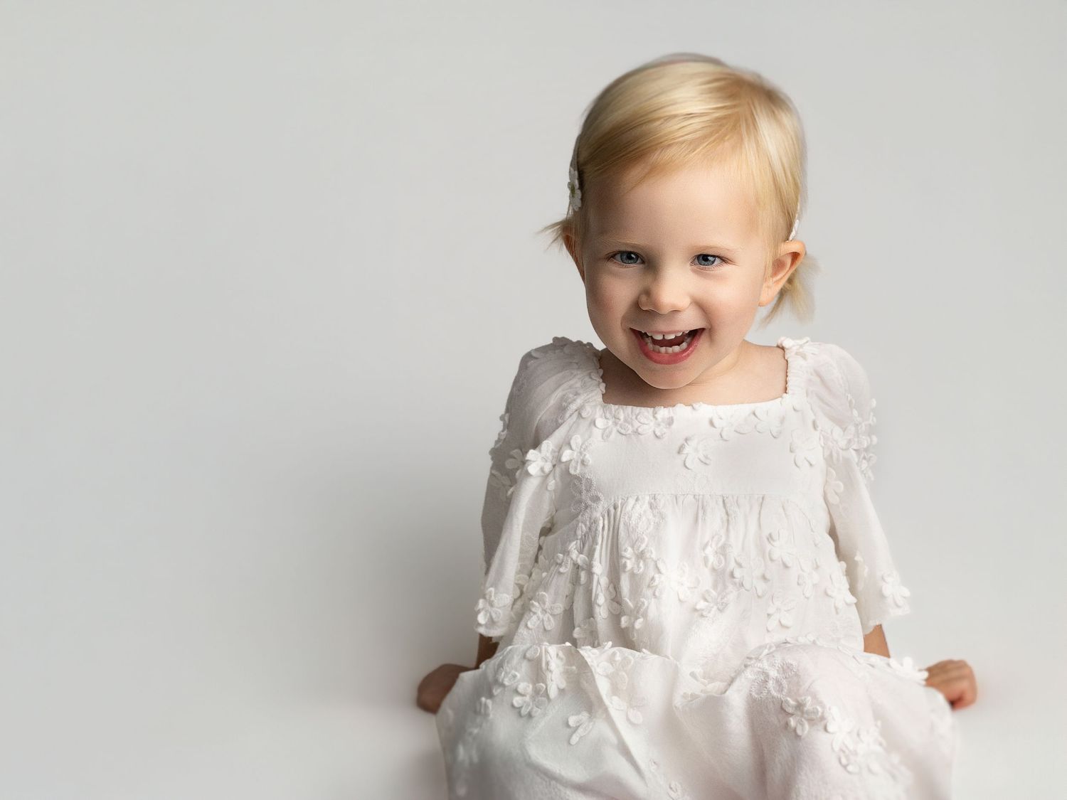 Smiling toddler girl in white lace floral dress with long sleeves, posed against light gray background.