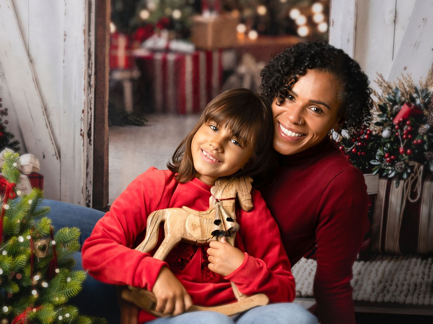 A woman and young girl in red sweaters pose together with a wooden reindeer toy in a festive Christmas setting.