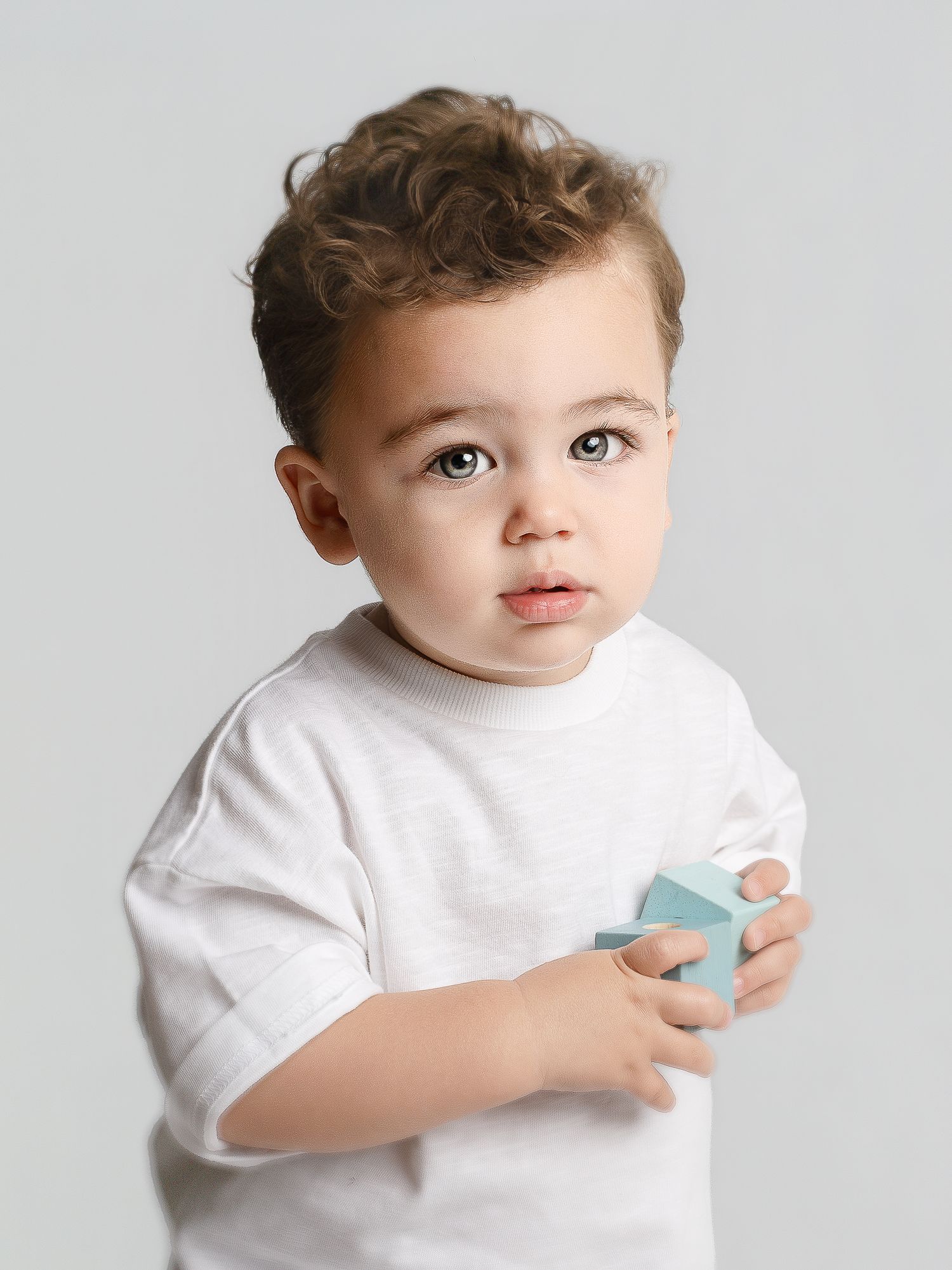 Cute toddler boy with curly hair holding a light blue block, wearing a white t-shirt against a gray background.