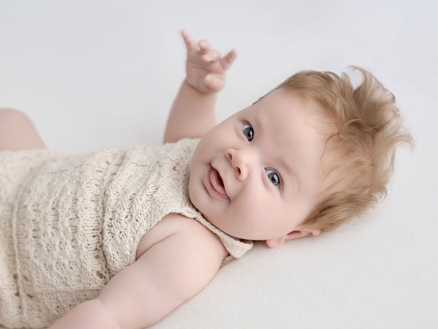 Adorable baby with blue eyes and light hair lying on white background, wearing a cozy knit romper, reaching up.