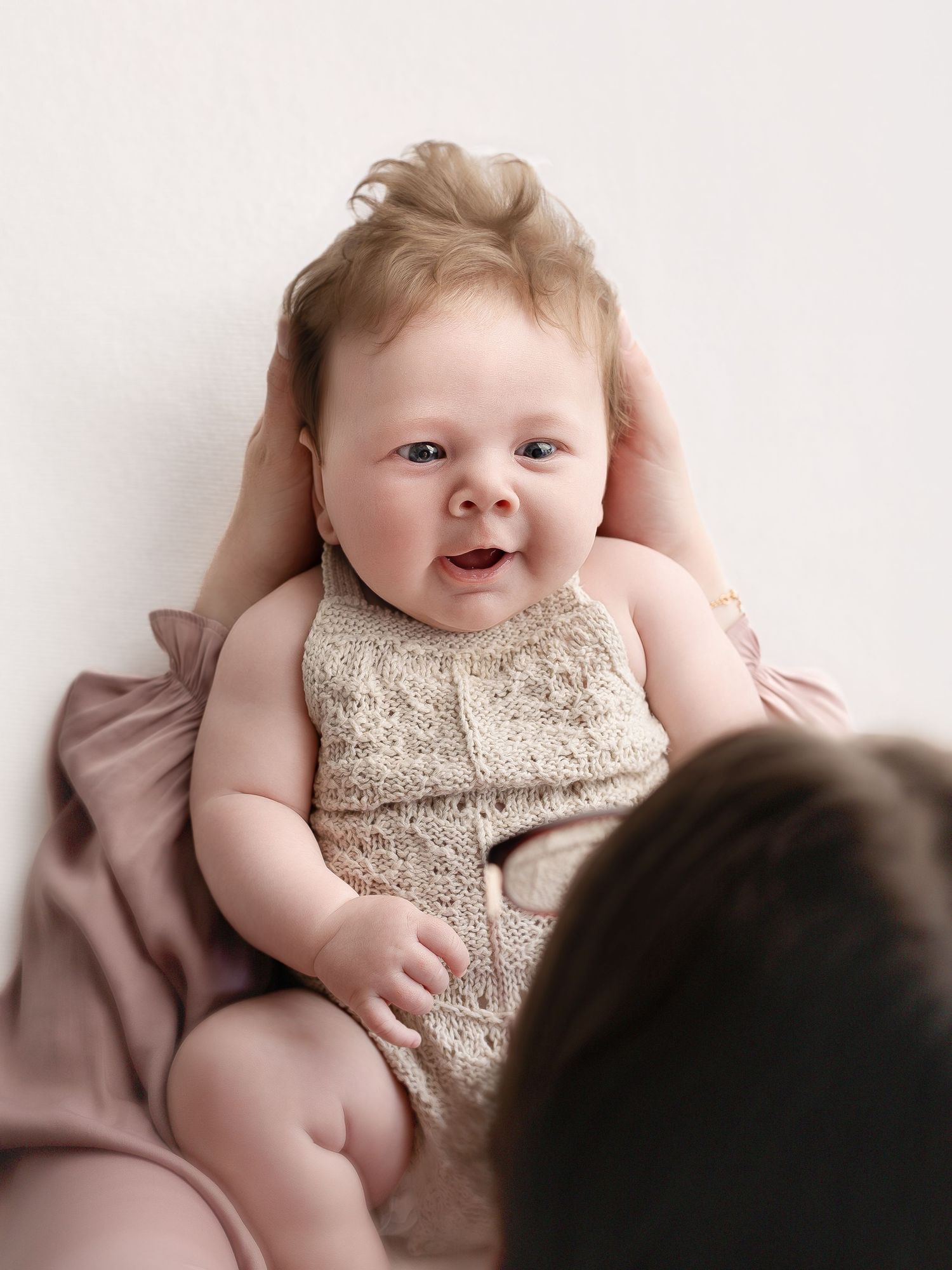 A smiling baby in a lace romper lies on a pink blanket, looking up at a person in a bright studio setting.