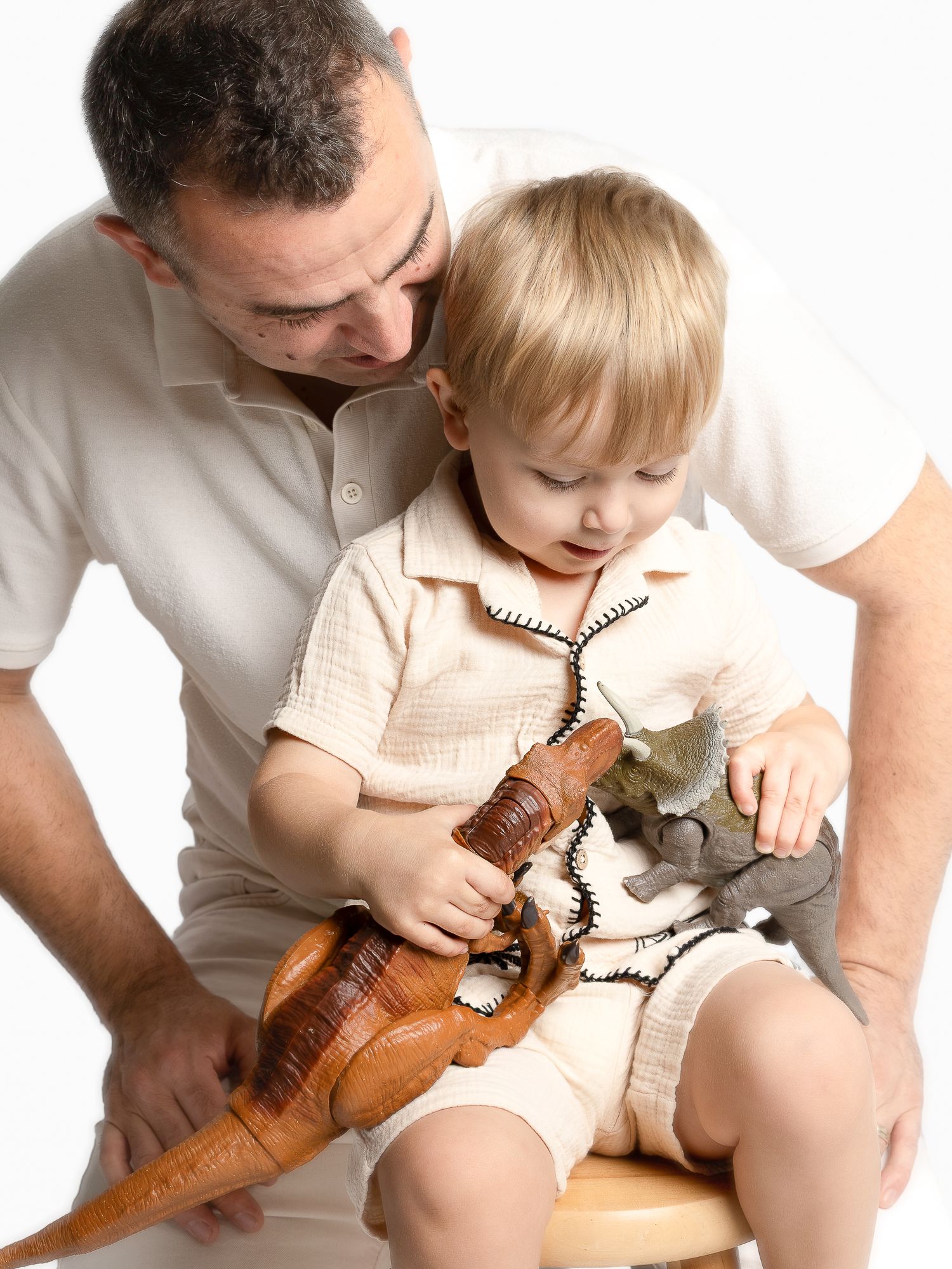 Father and toddler son playing with toy dinosaurs together while sitting on a stool against white background.