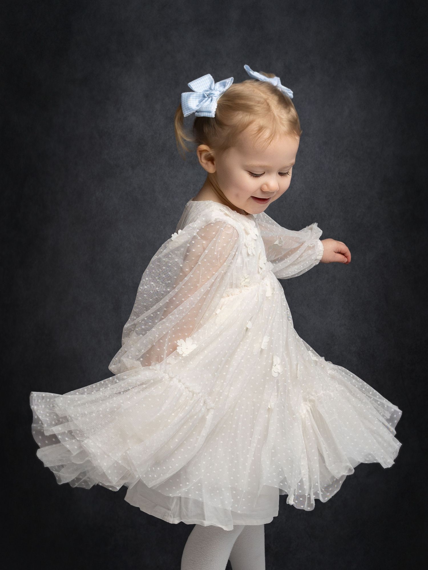 Toddler girl in white tulle dress with bow hair clip twirling and smiling against dark backdrop.