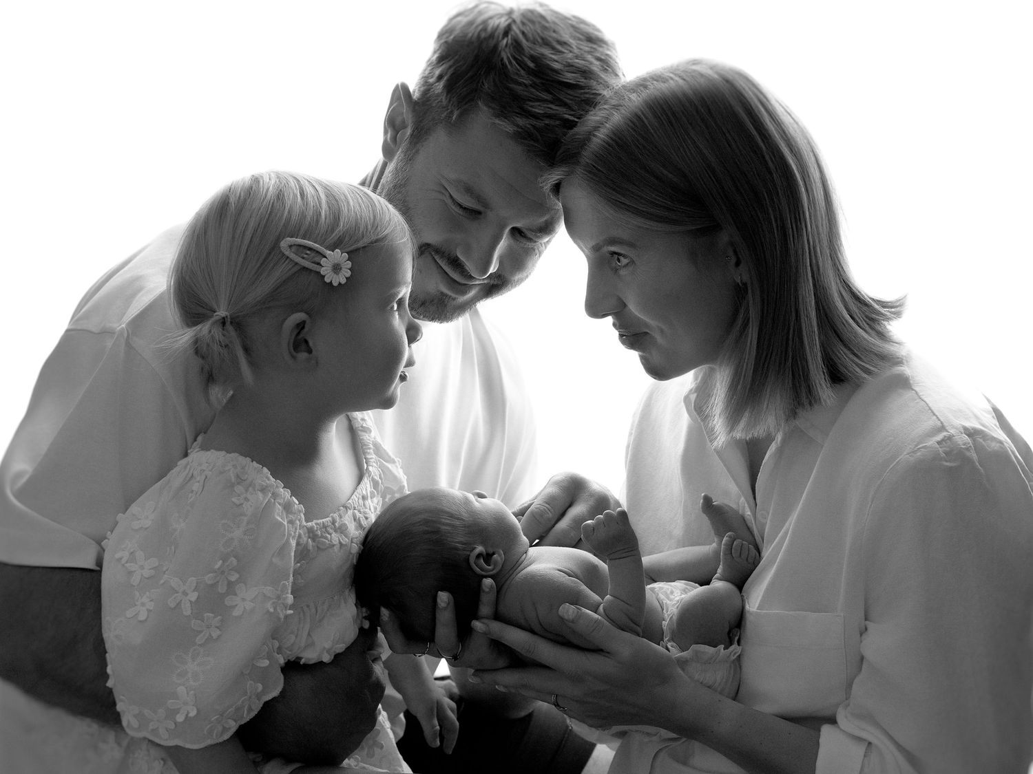 Black and white family portrait with parents and toddler girl lovingly gazing at newborn baby.