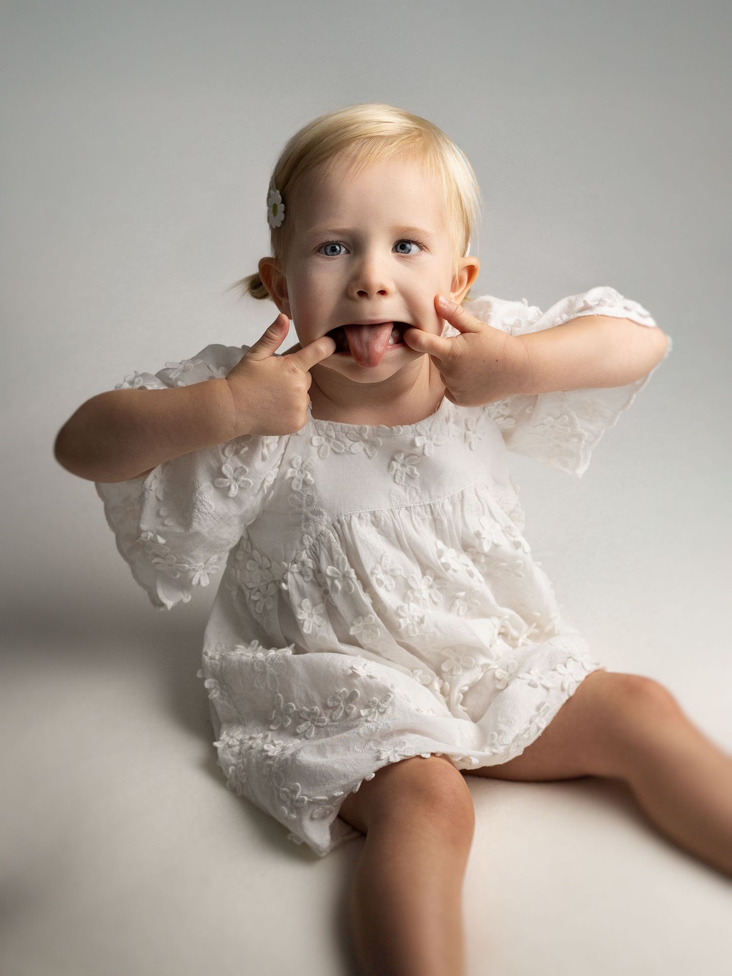 Playful child in white dress making silly faces and poses on grey backdrop.