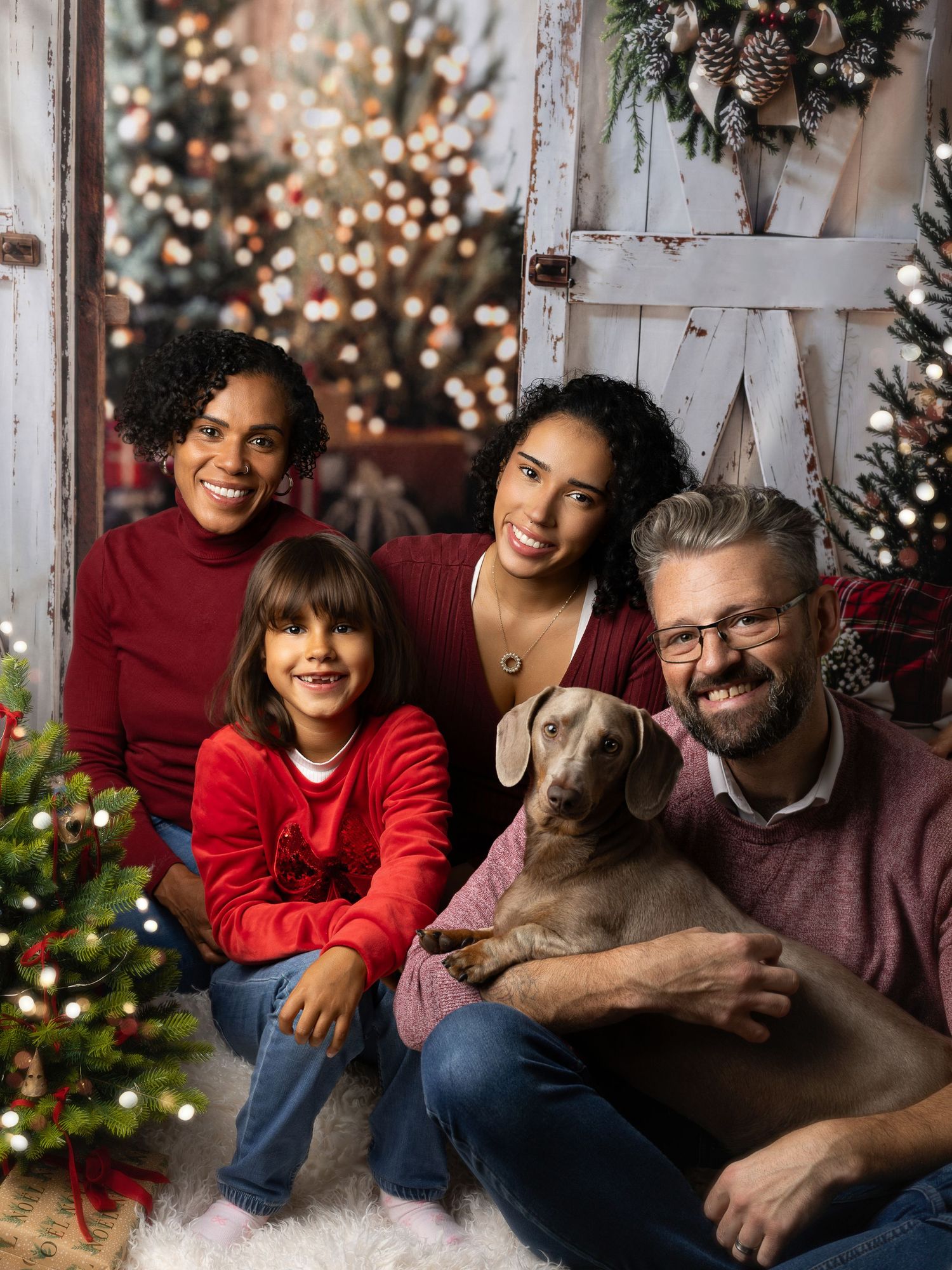 A joyful family of four poses with their Weimaraner dog in front of festive Christmas decorations and twinkling lights.