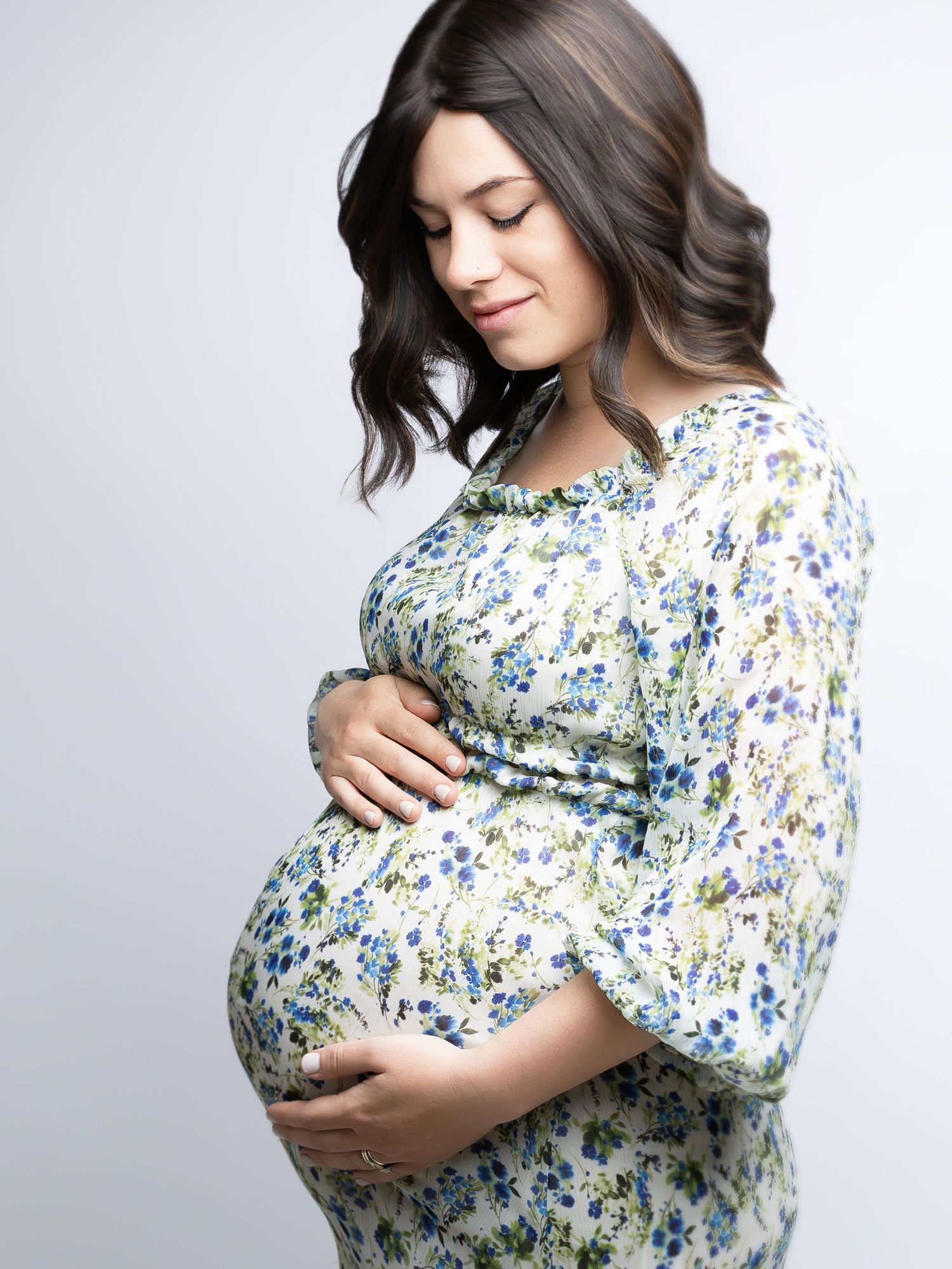 Pregnant woman in floral dress cradles baby bump against light background in maternity photoshoot series.