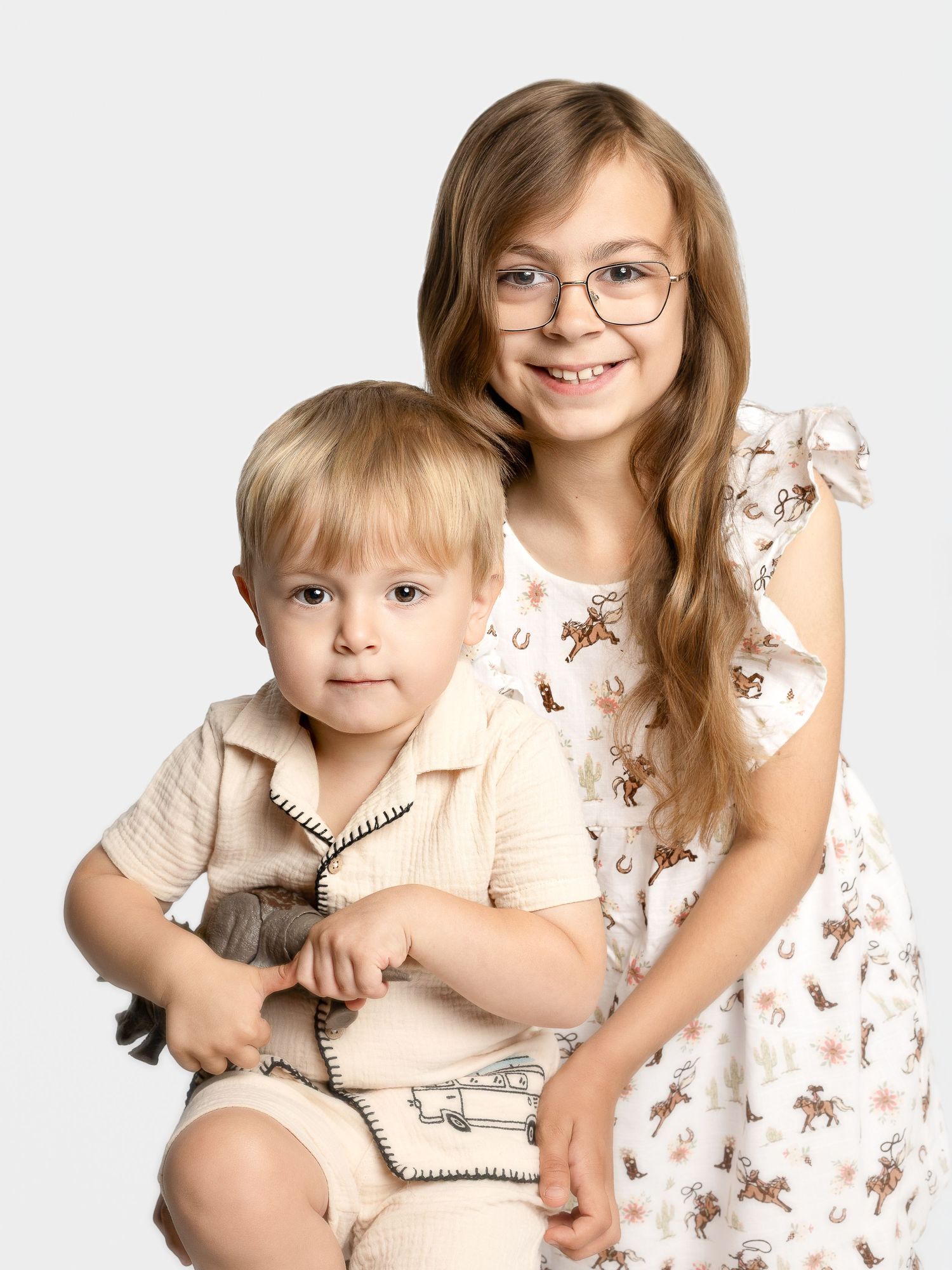 Two young siblings pose together, older girl with glasses in floral dress hugging toddler boy in cream outfit against white background.