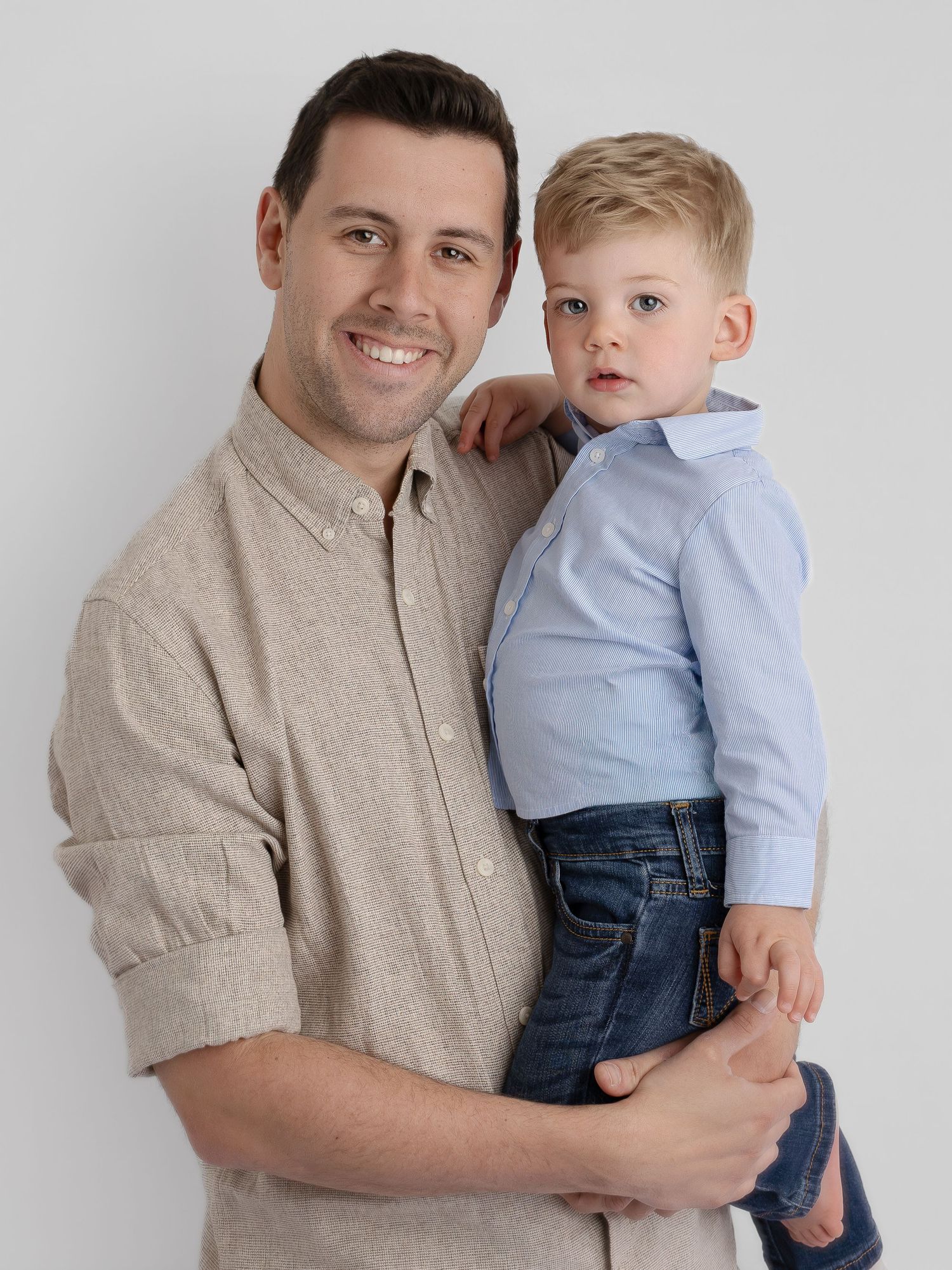A smiling man in a beige shirt holds a young blonde boy in a blue shirt and jeans against a white background.