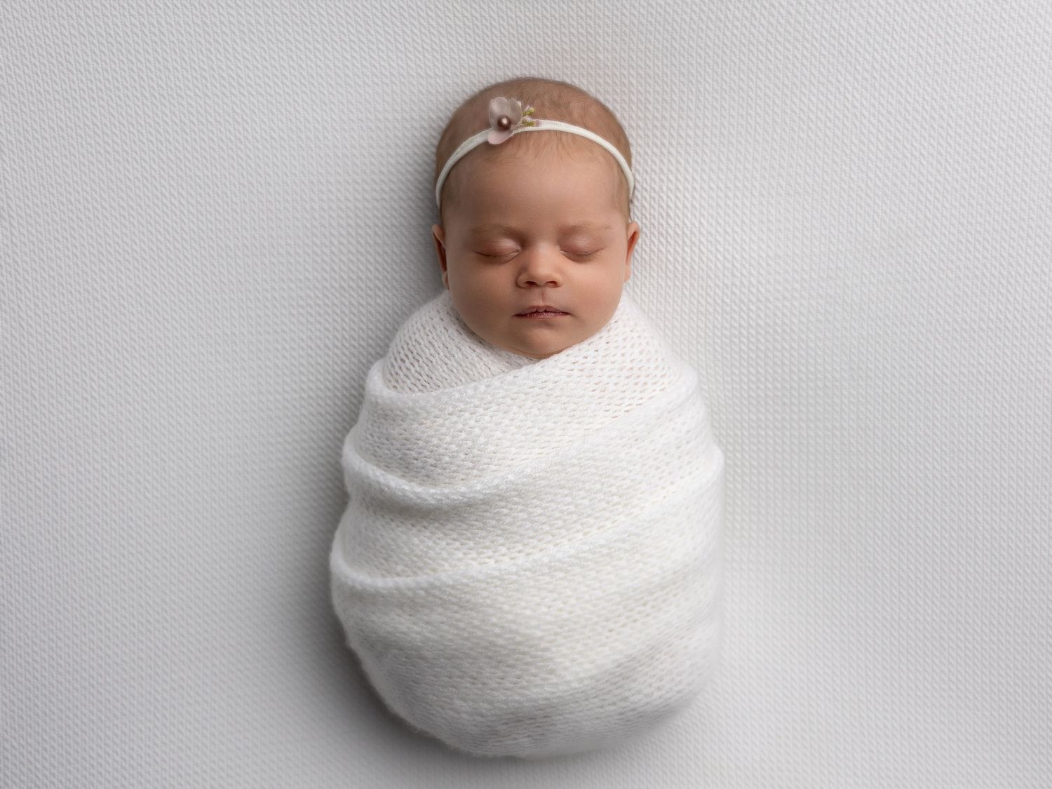 Peaceful newborn baby wrapped in white swaddle wearing delicate headband against grey background.