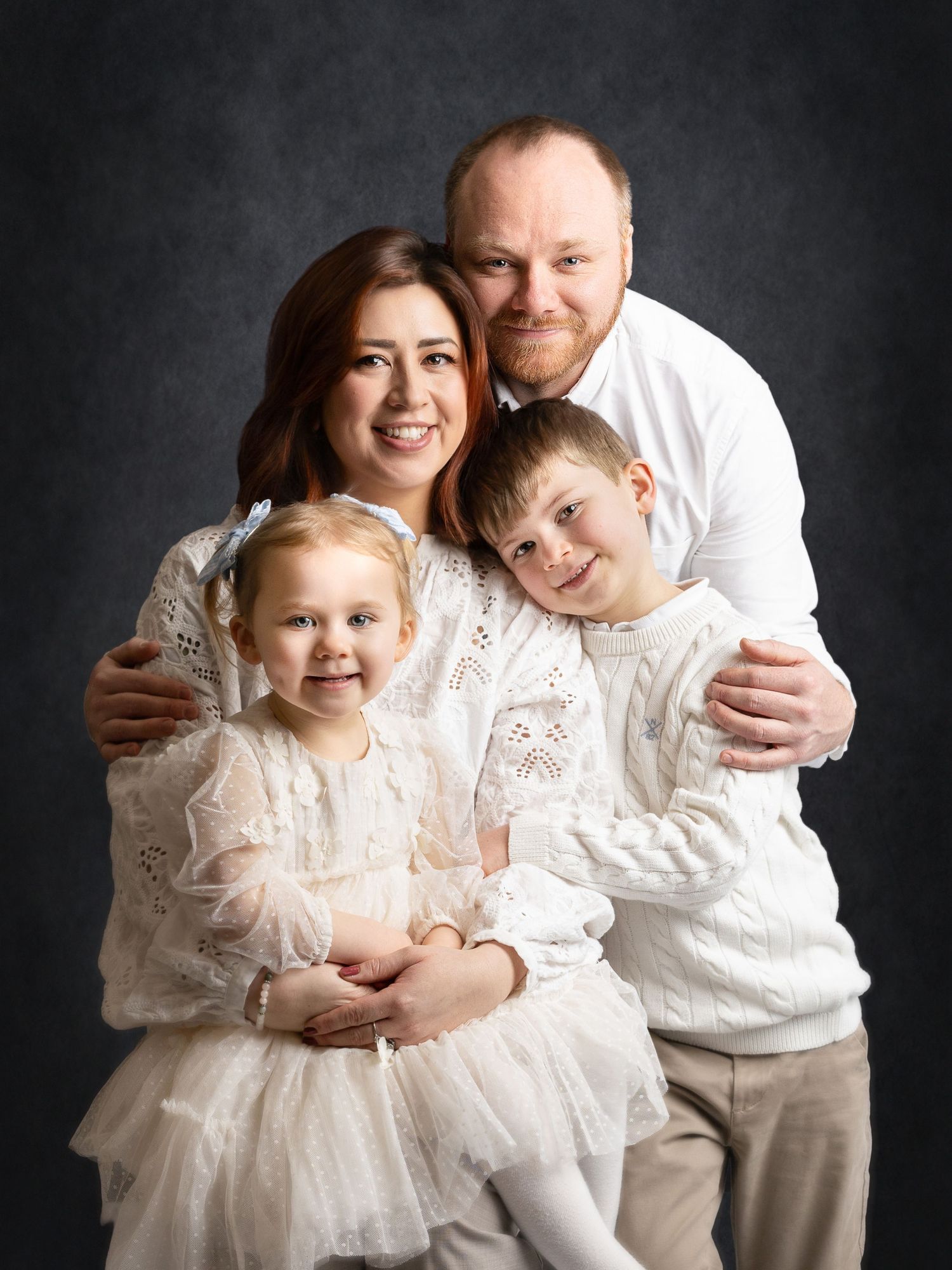 A smiling family of four poses in white outfits against a dark backdrop in a professional studio portrait.