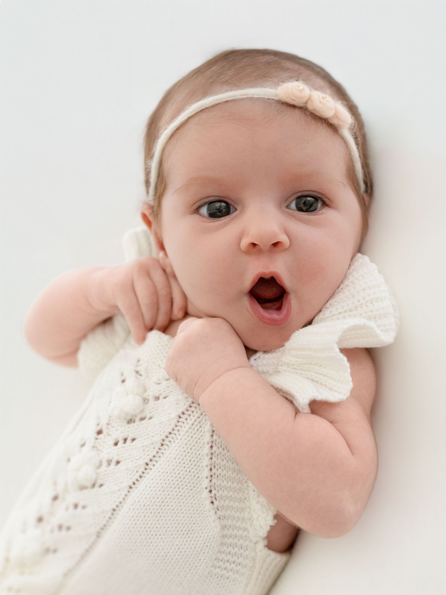 A wide-eyed newborn baby girl in a white knit outfit and floral headband lies on a white background with mouth open.