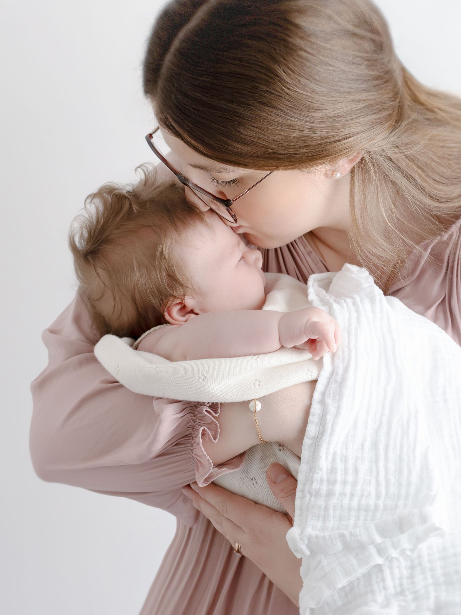A mother wearing glasses kisses her sleeping newborn baby dressed in white, against a soft white background.