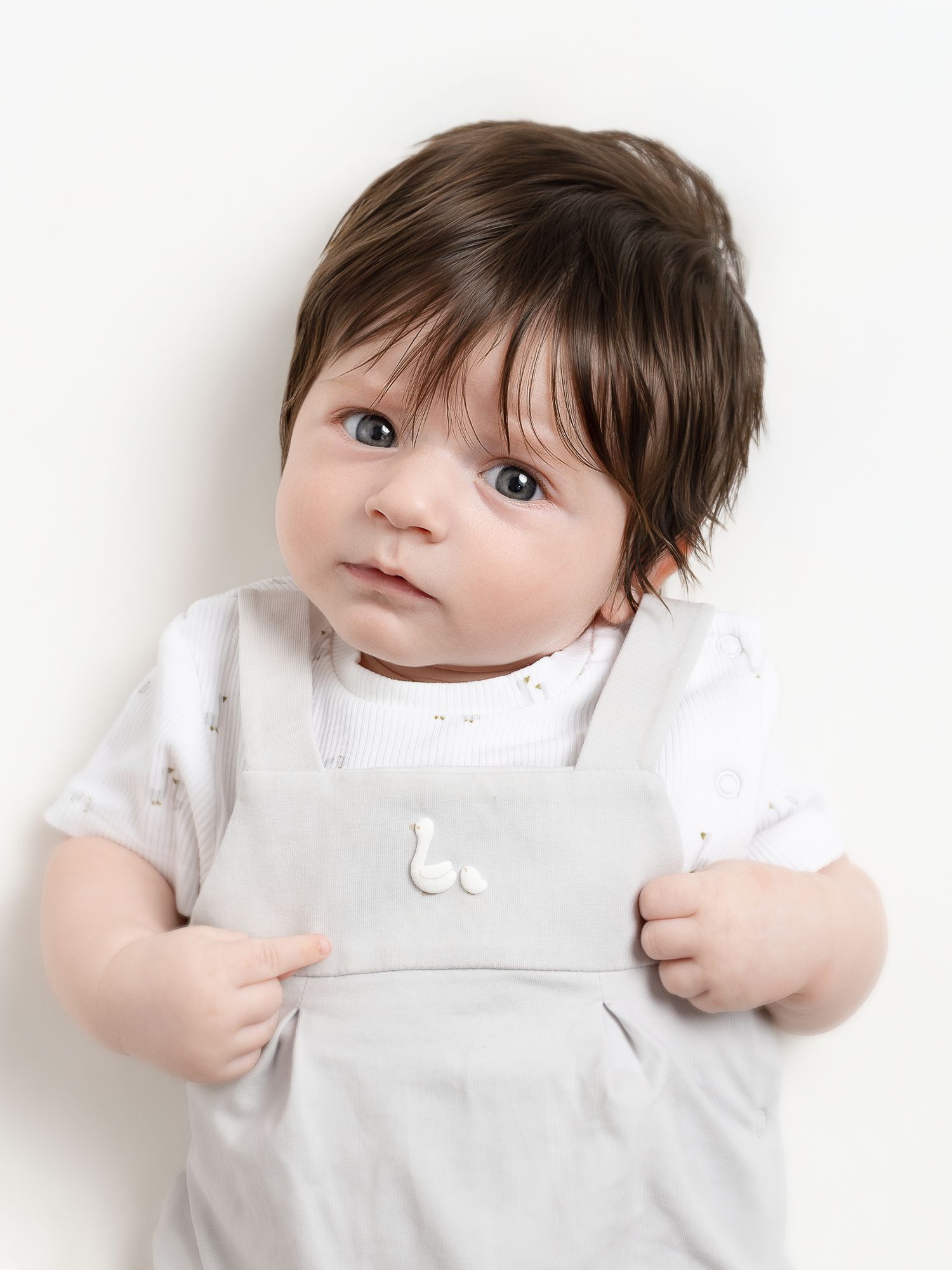 Adorable baby with dark hair wearing a white onesie and gray overalls with duck buttons on a white background.
