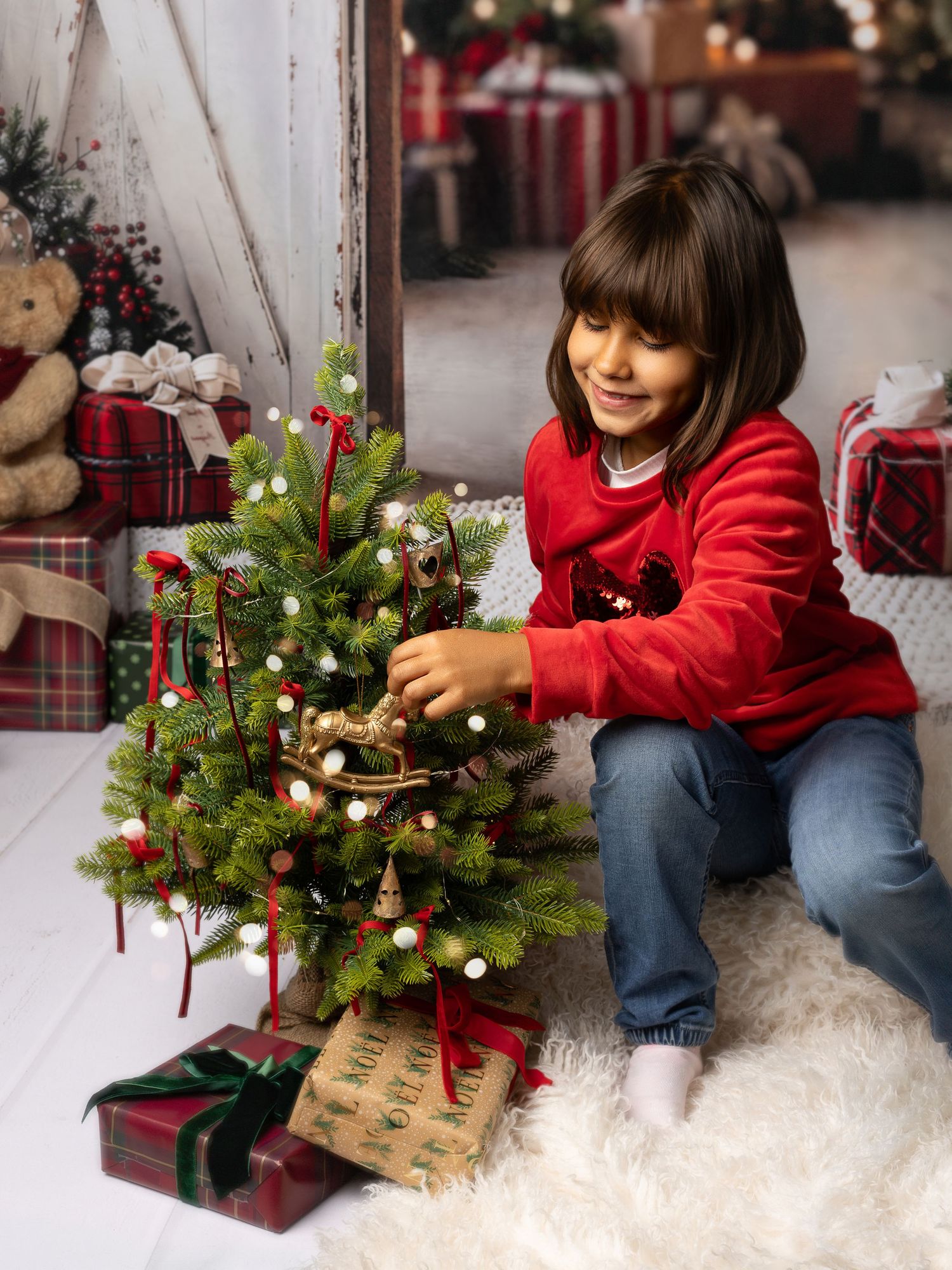 A smiling girl in a red sweater decorates a small Christmas tree surrounded by wrapped gifts on a white fur rug.
