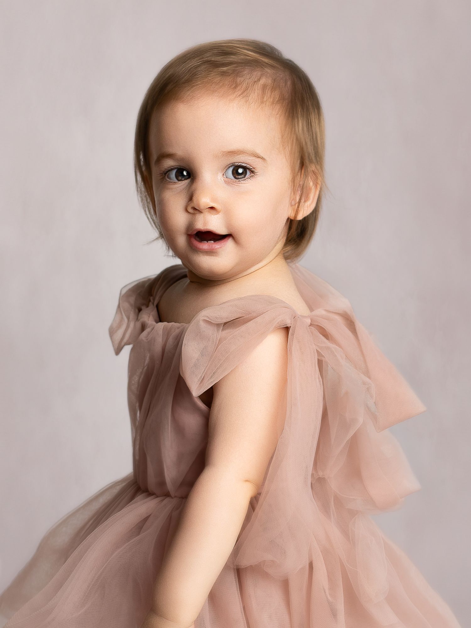 Toddler girl in a blush pink tulle dress with bow details smiles over her shoulder in a studio portrait.