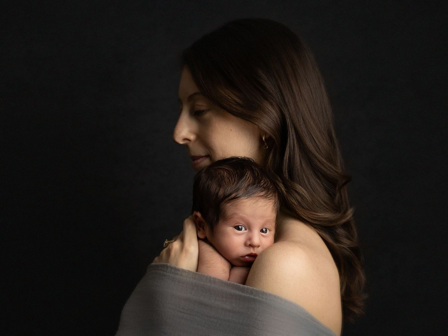 Intimate mother and newborn portrait against dark background captures tender moment.