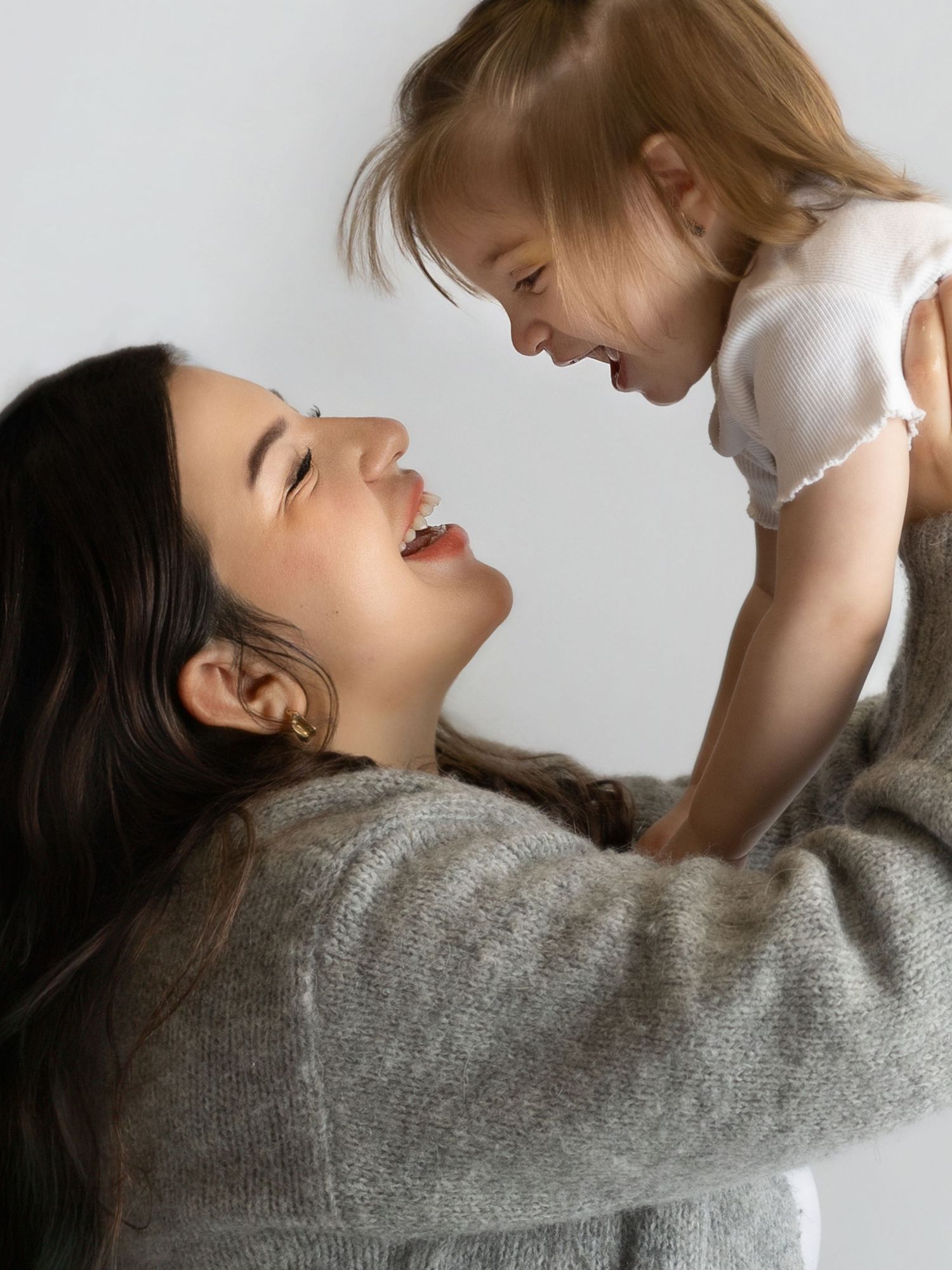 A parent in a gray sweater lifts and plays with a joyful toddler wearing white against a light background.