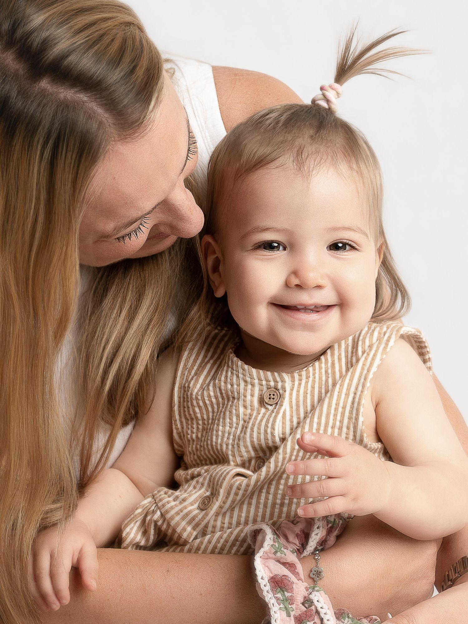 A smiling baby girl in a striped dress being held by a blonde woman in a white studio setting.