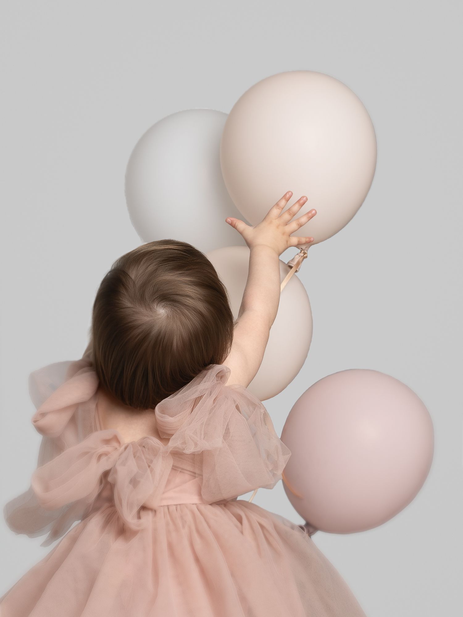 A toddler in a pink tulle dress reaches toward pastel pink and white balloons against a gray background.