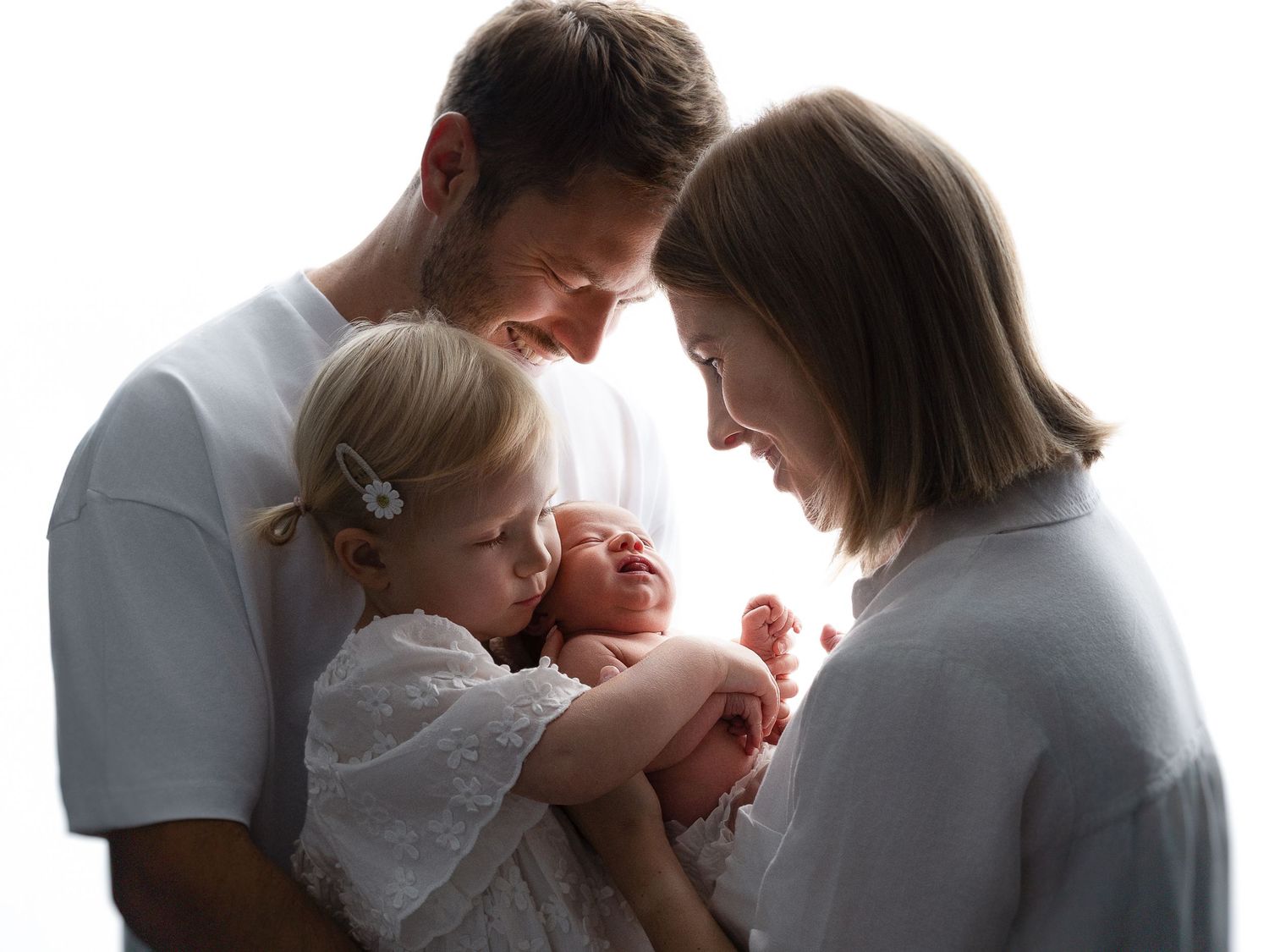 Family of four with parents in white shirts tenderly gazing at newborn baby held by young toddler daughter.