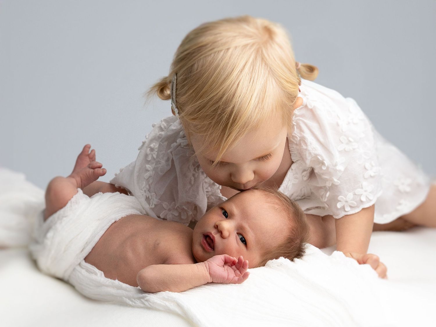 Sweet moment between siblings as older child in white lace dress kisses newborn baby.