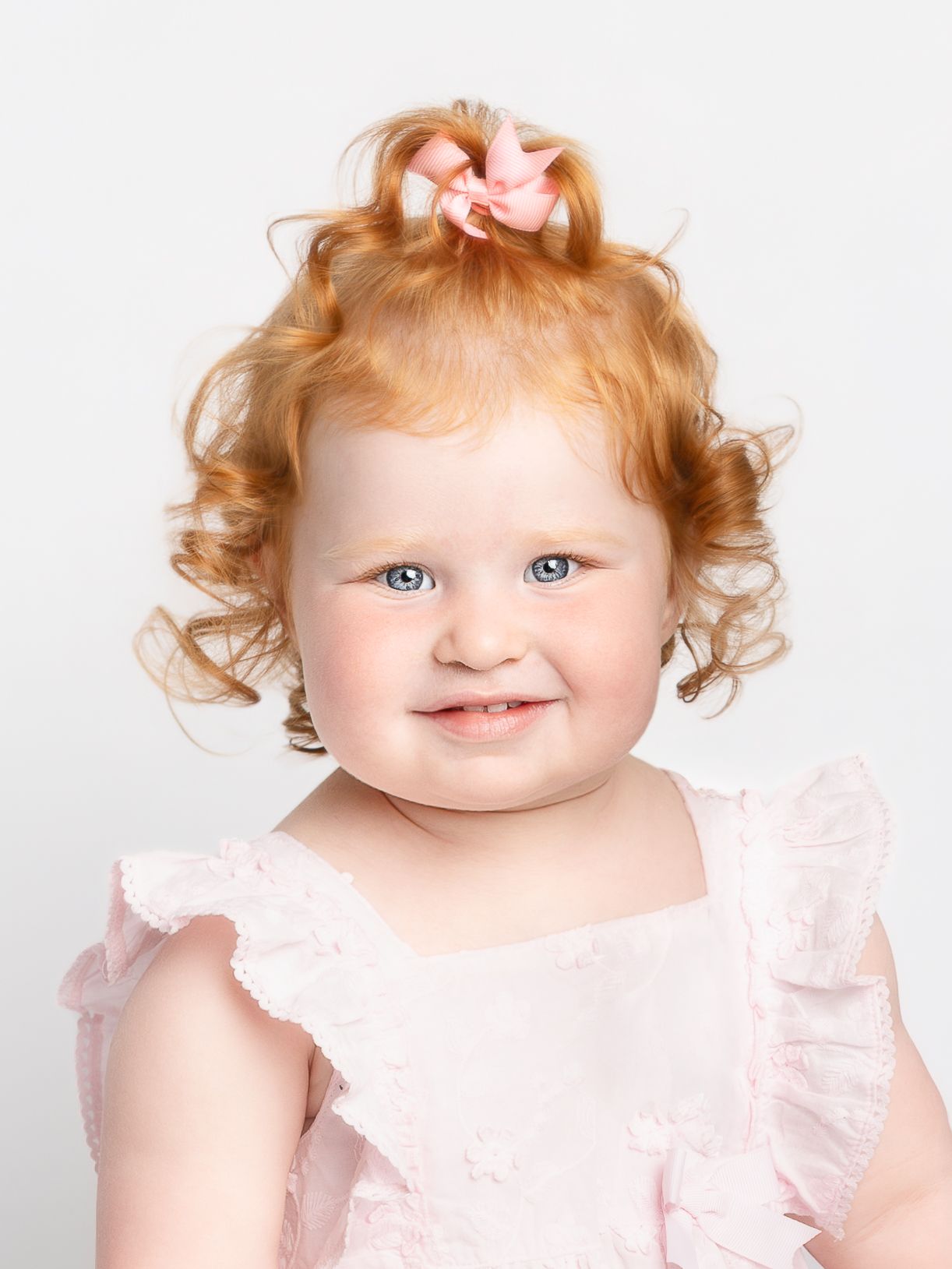 Adorable toddler girl with curly red hair and blue eyes wearing a pink ruffled dress with a pink bow.
